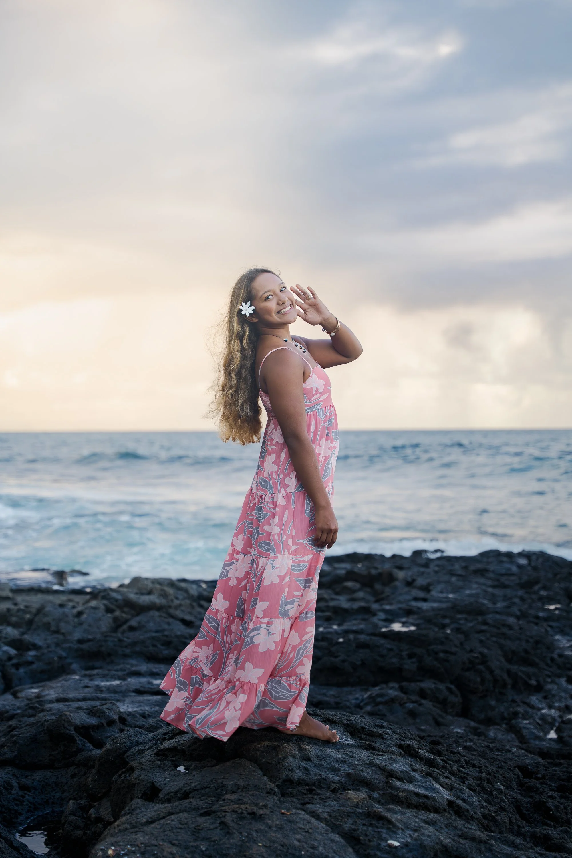 A woman in a pink floral dress standing on black volcanic rocks by the ocean, smiling and waving at the camera with a flower in her hair, during sunset.