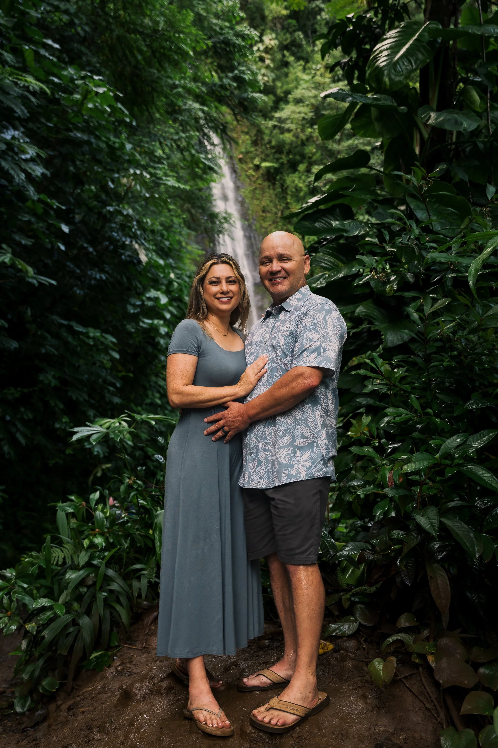 A couple stands together in a lush green jungle with a waterfall in the background. The woman wears a gray dress and sandals, and the man wears a patterned shirt, shorts, and sandals. They are smiling and embracing each other.