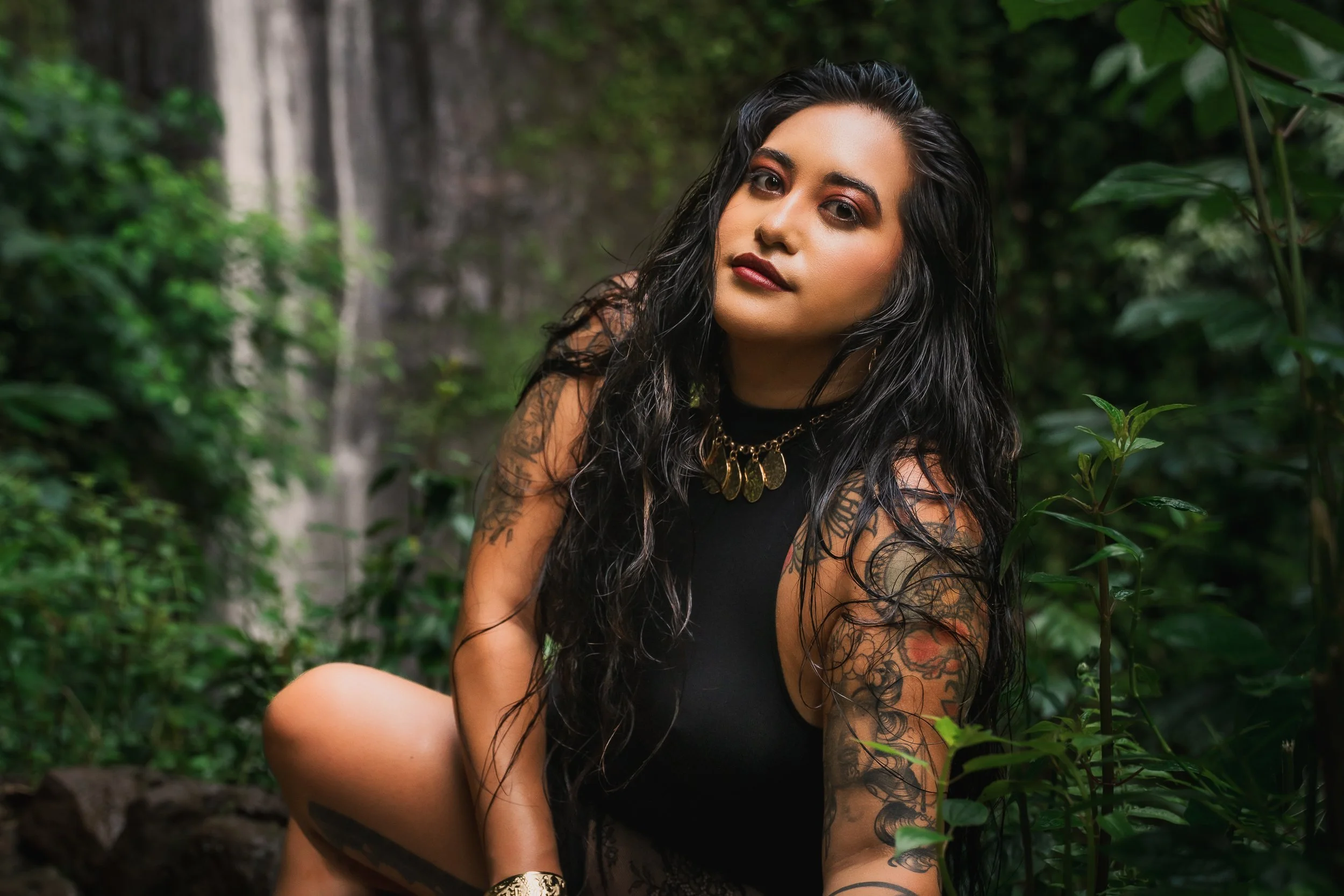 A woman with long black hair and tattoos wearing a black sleeveless top, sitting in a lush green forest with a waterfall in the background.