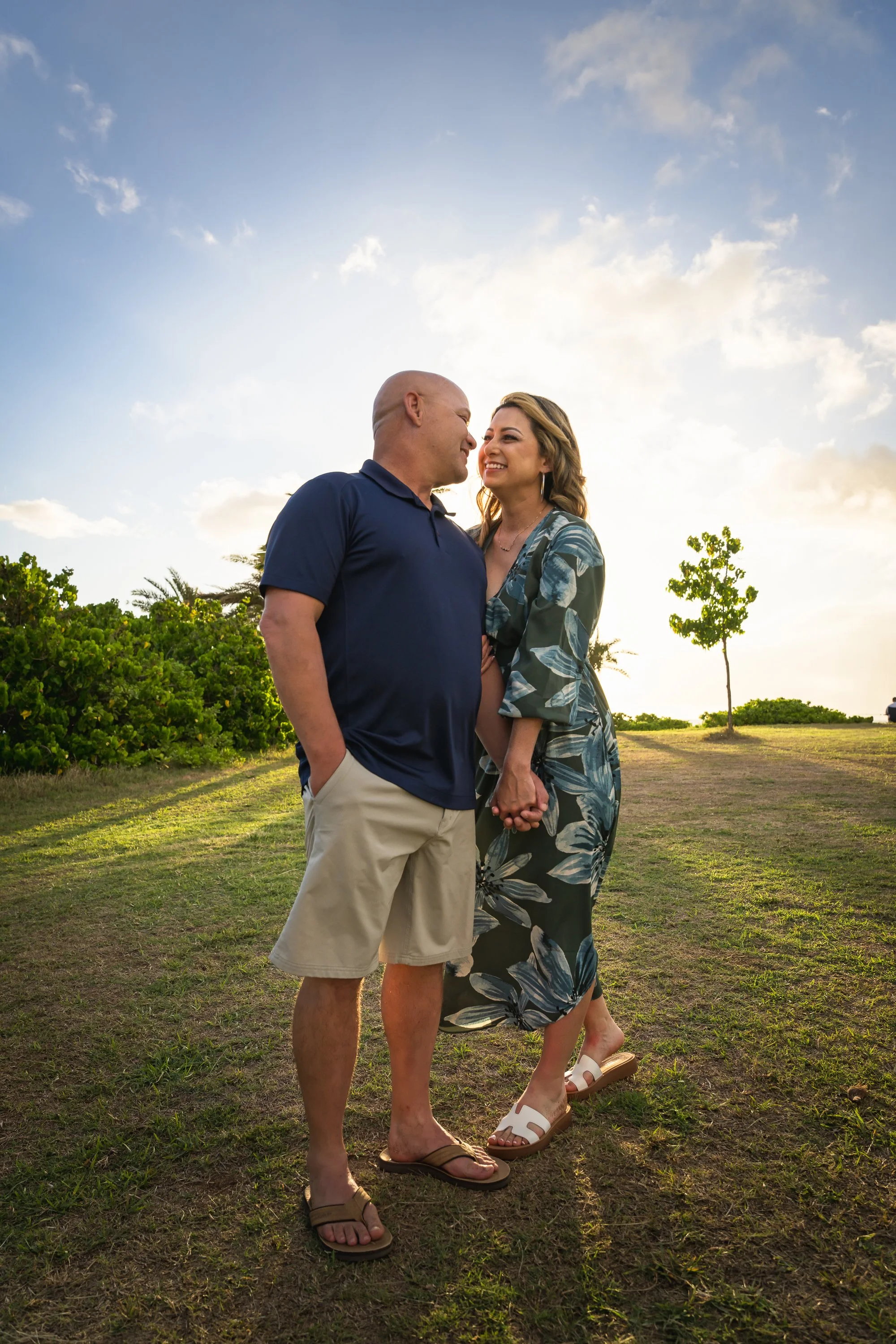 A couple holding hands and smiling at each other outdoors during sunset with a grassy field and trees in the background.