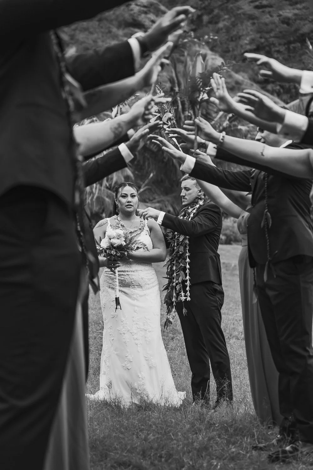 Black and white photograph of a wedding ceremony outdoors with a bride in a white dress holding a bouquet, and a groom wearing a suit with a floral lei, surrounded by guests with outstretched hands forming an arch.