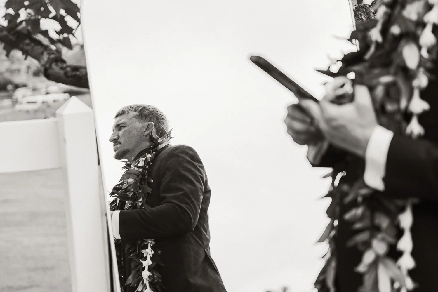 A man dressed in formal attire and wearing a lei, standing outdoors with a reflection visible in a mirror or glass, during daylight. Another person with a lei is in the foreground using a smartphone.