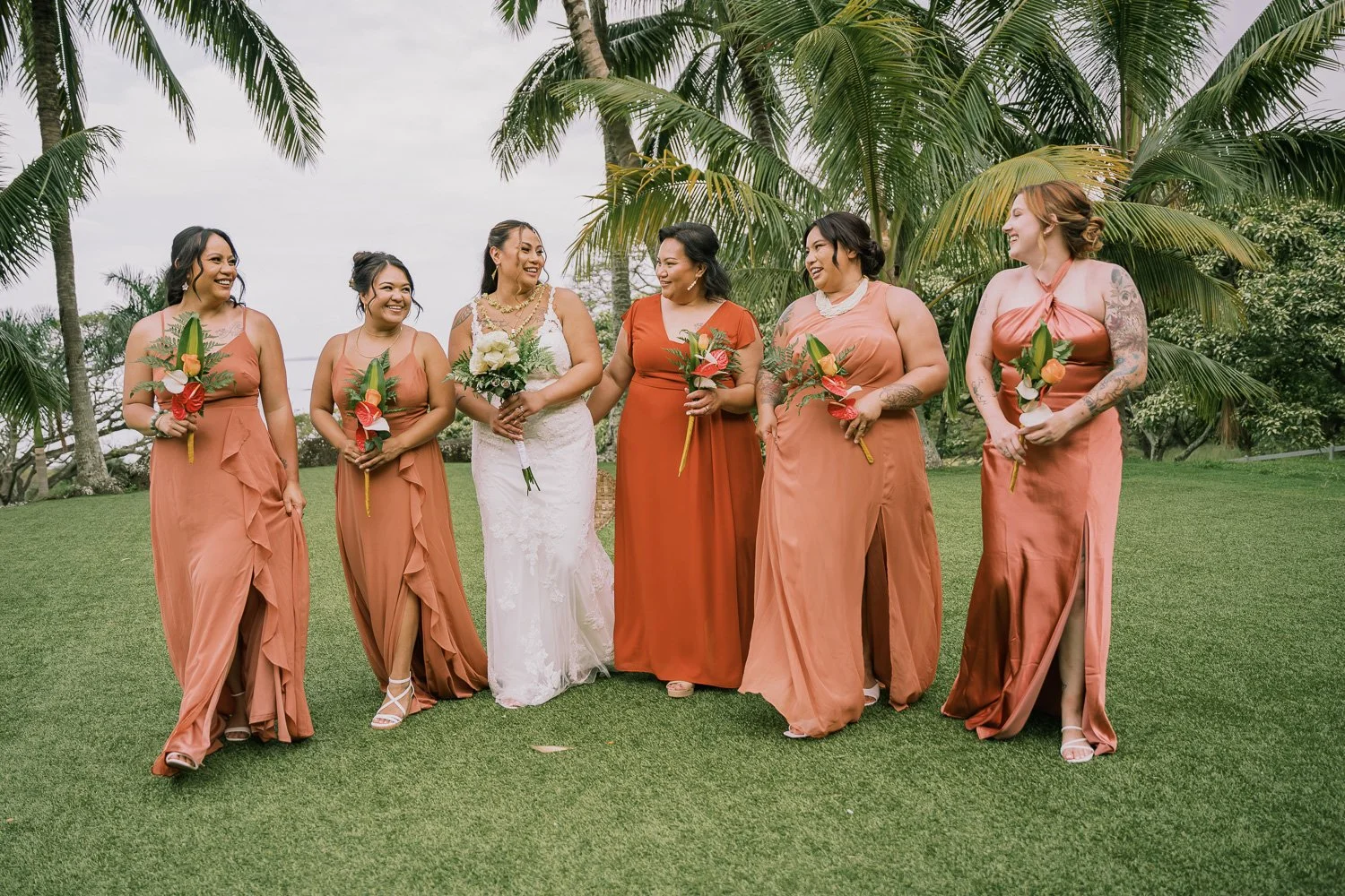 Bridal party with six women standing on grass, with palm trees in background, smiling and holding bouquets, during a wedding celebration.