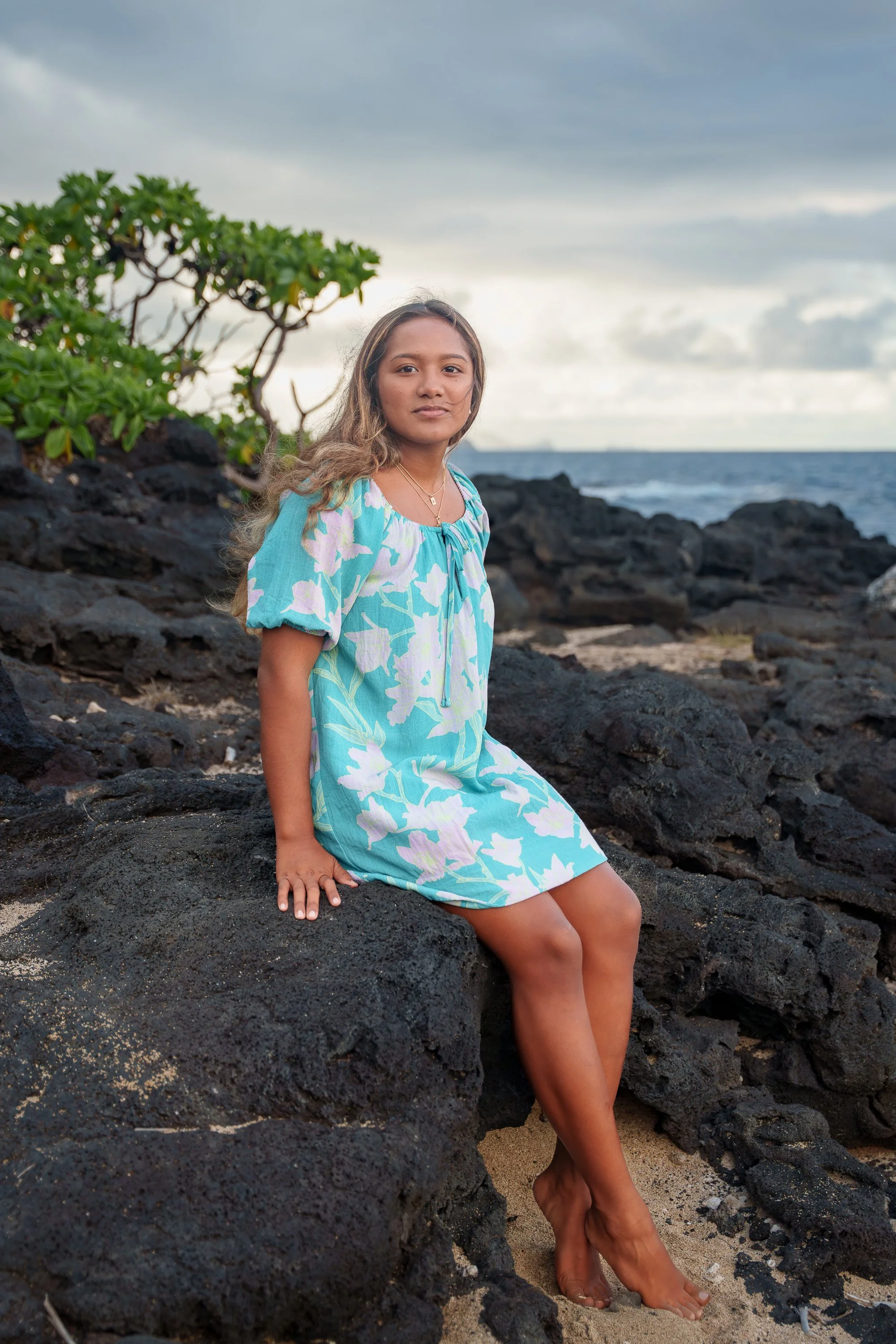 A young girl with long, wavy hair sitting on black volcanic rocks along a beach under a cloudy sky, wearing a light blue dress with white floral patterns.