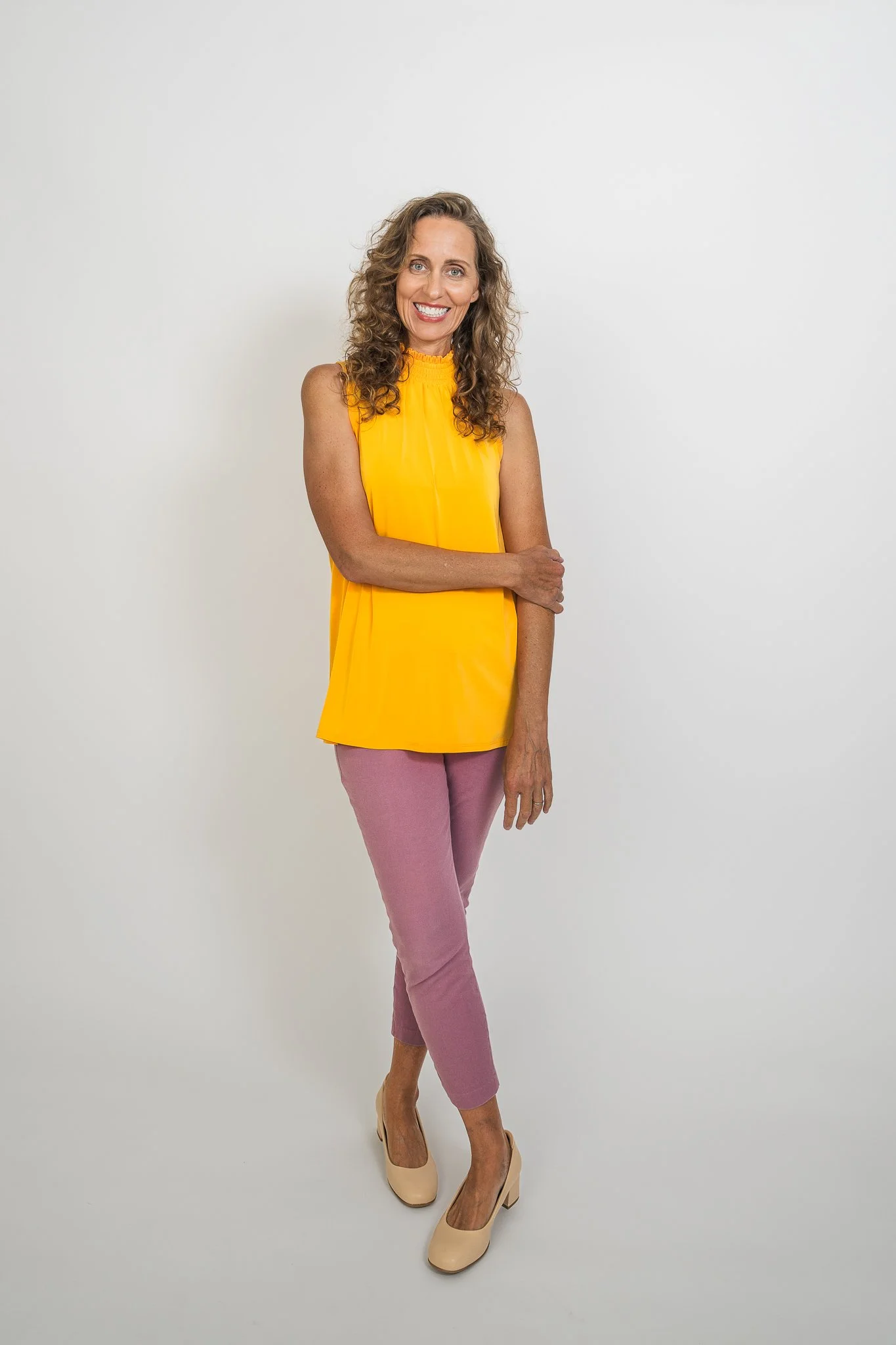 A woman with curly hair wearing a yellow sleeveless top, pink pants, and beige heels, smiling and standing against a plain white background.