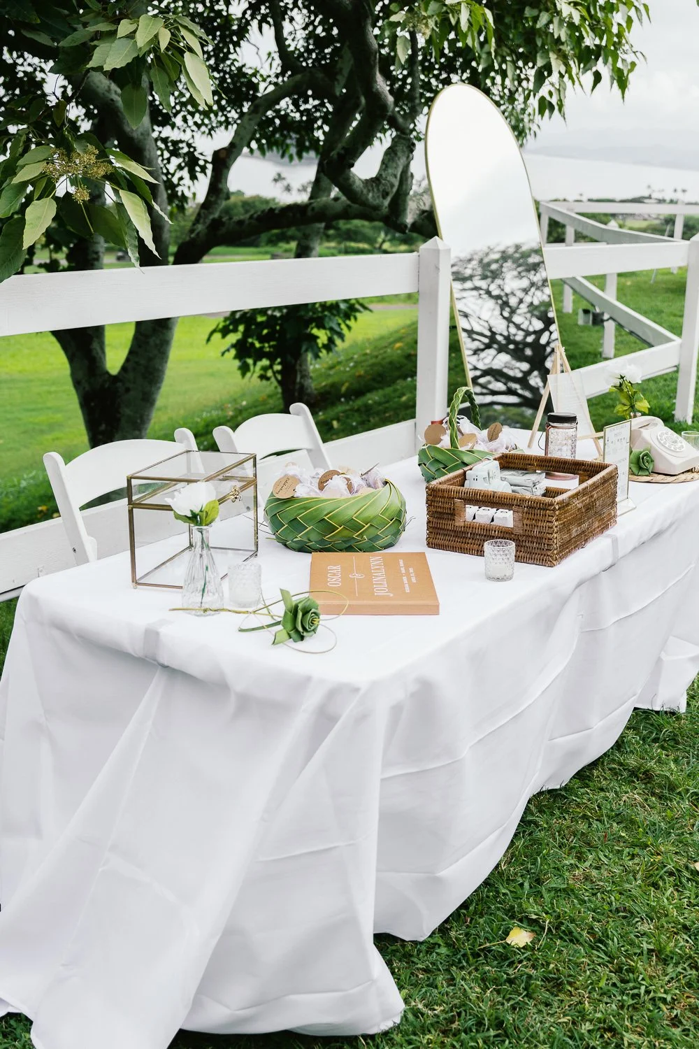 Outdoor table decorated with a white tablecloth, green leaves, and decorative objects, set in a lush garden with trees and water in the background.