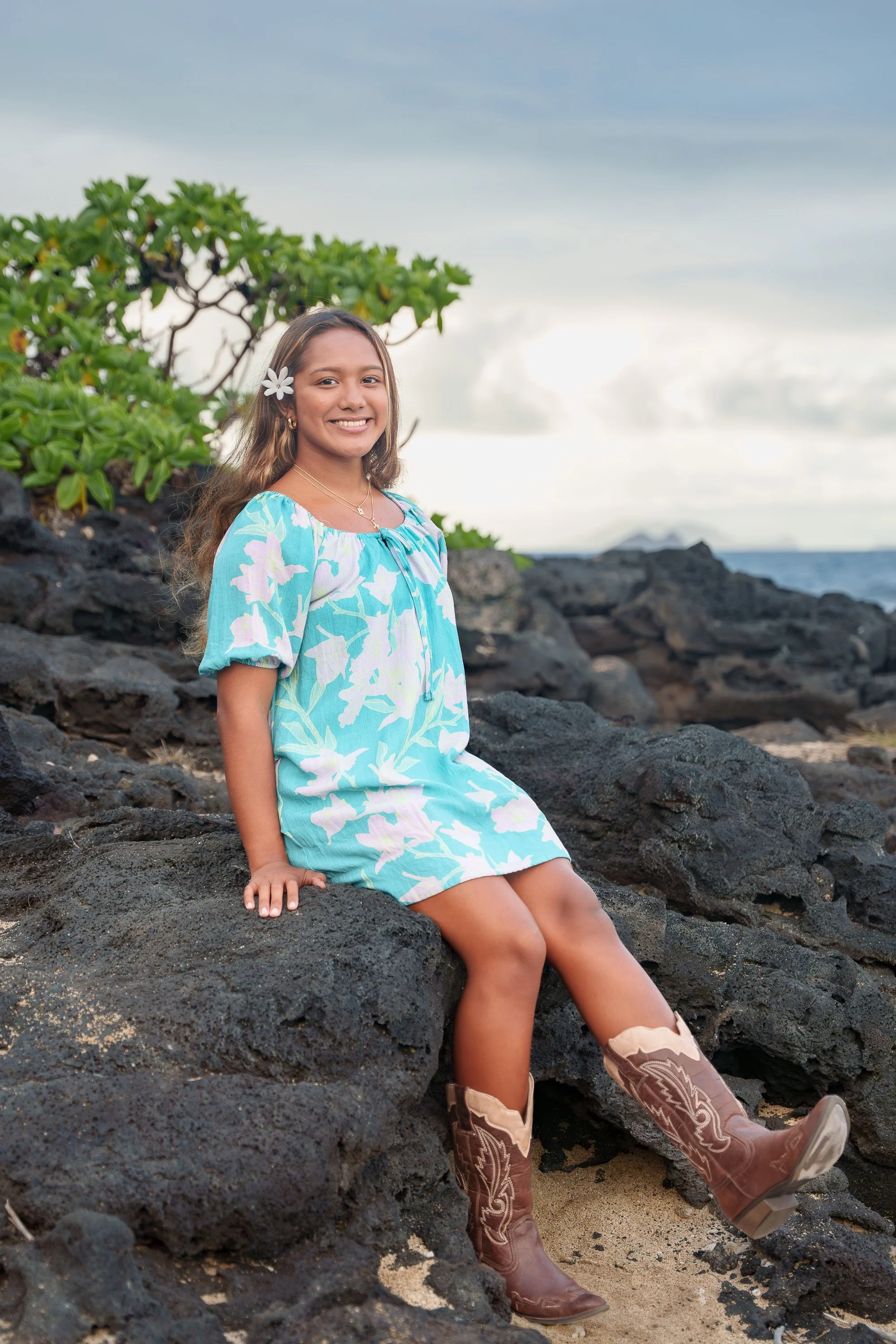 A young girl sitting on black volcanic rocks at the beach, wearing a blue and white floral dress, cowboy boots, and a white flower in her hair, smiling with green foliage and cloudy sky in the background.