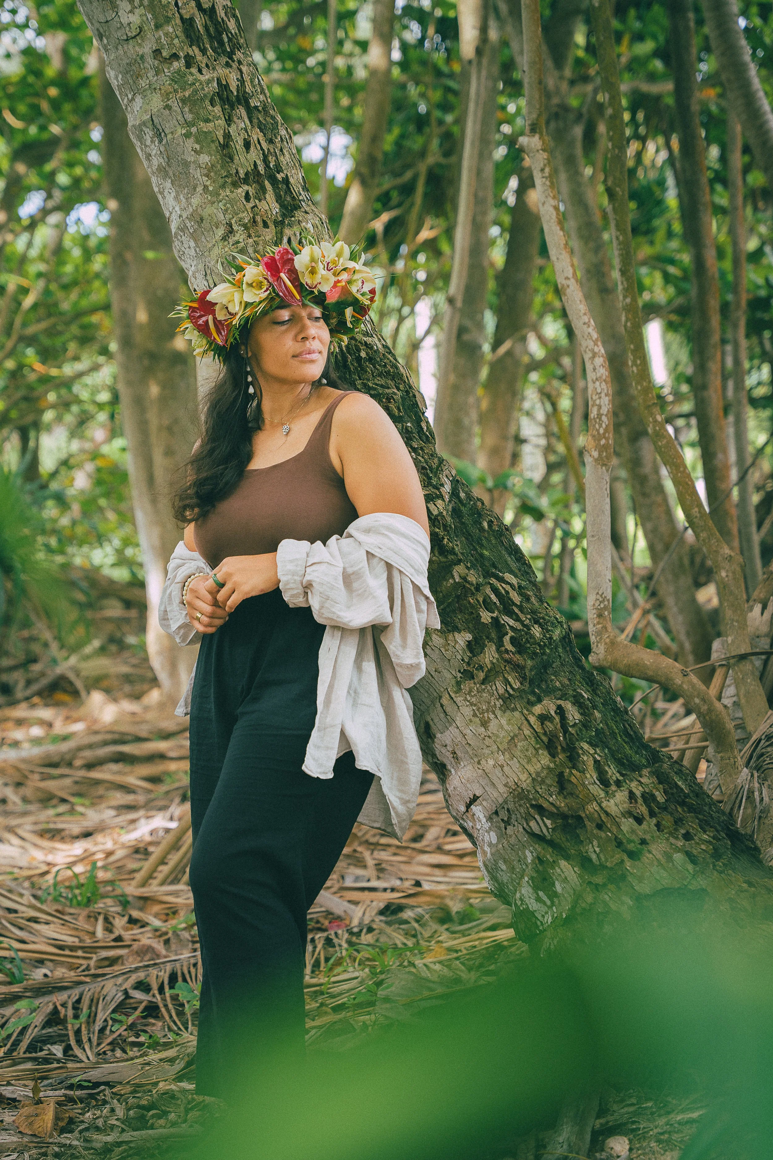 A woman with long dark hair wearing a floral crown, brown tank top, and black pants, standing in a lush, green forest, leaning against a tree with her eyes closed.