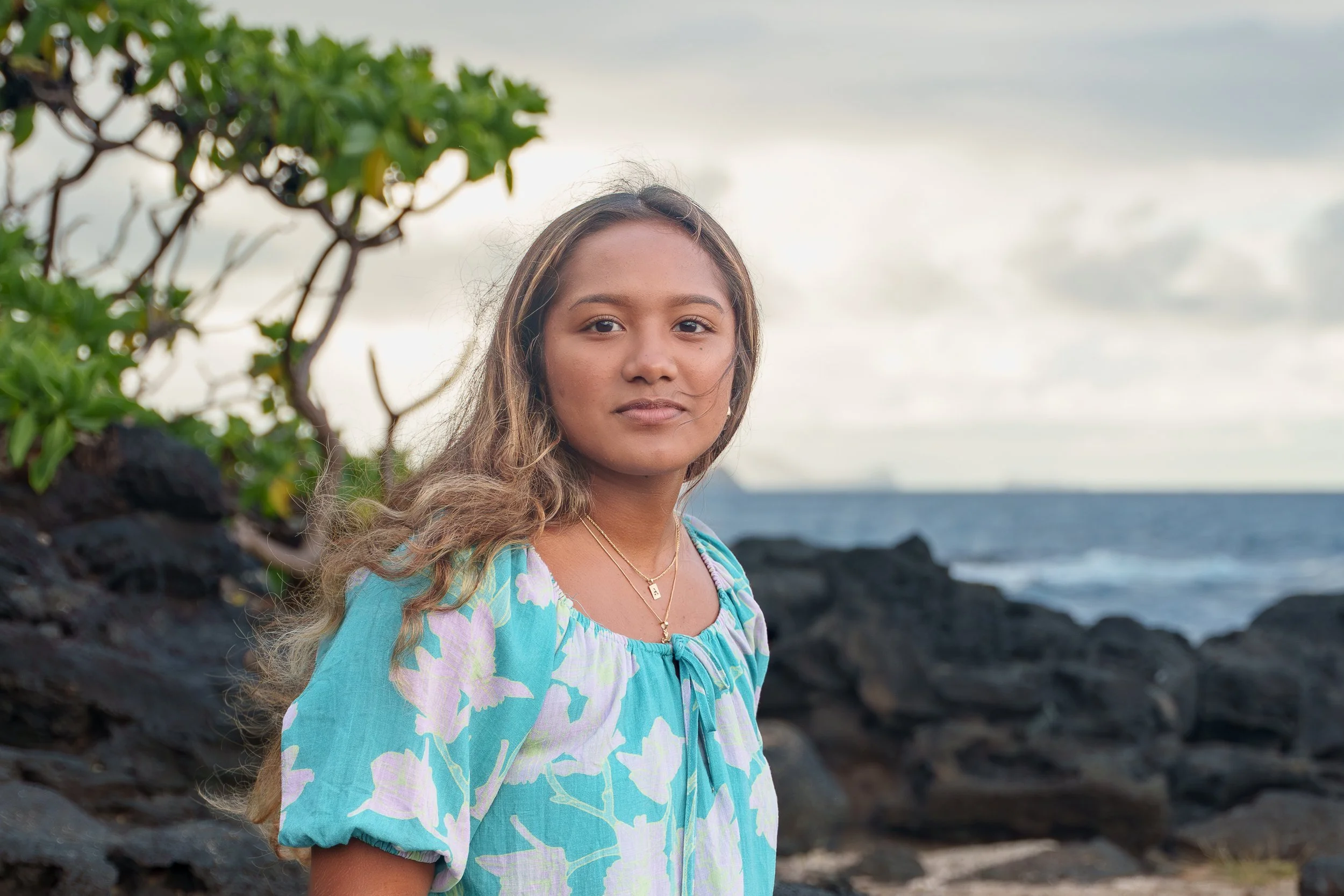 A young woman stands on a rocky beach with the ocean in the background, wearing a light blue floral dress and gold jewelry, looking at the camera.