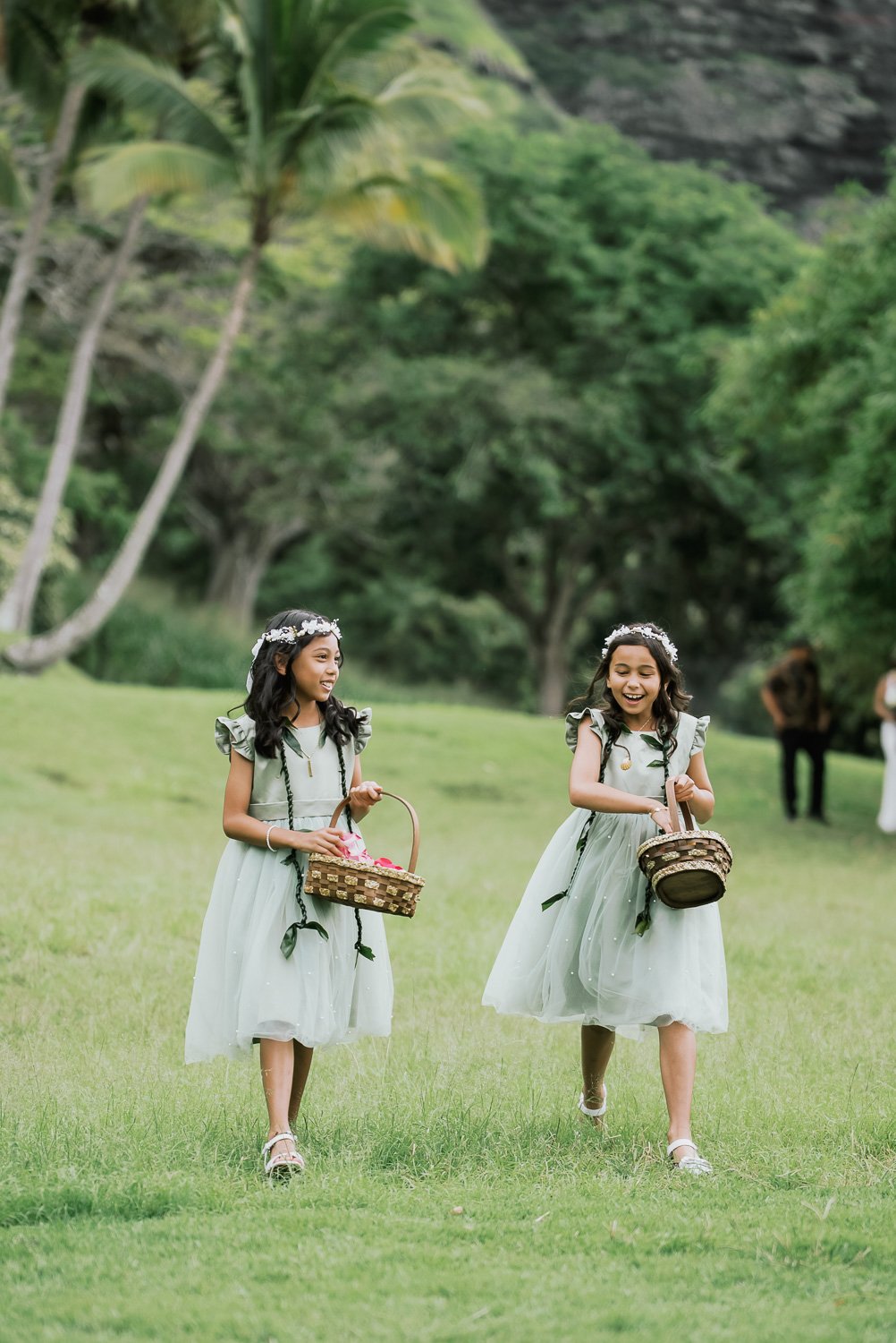 Two young girls in matching light green dresses with floral headbands carrying baskets, walking on a grassy area outdoors, surrounded by trees and greenery, likely participating in a celebration or event.