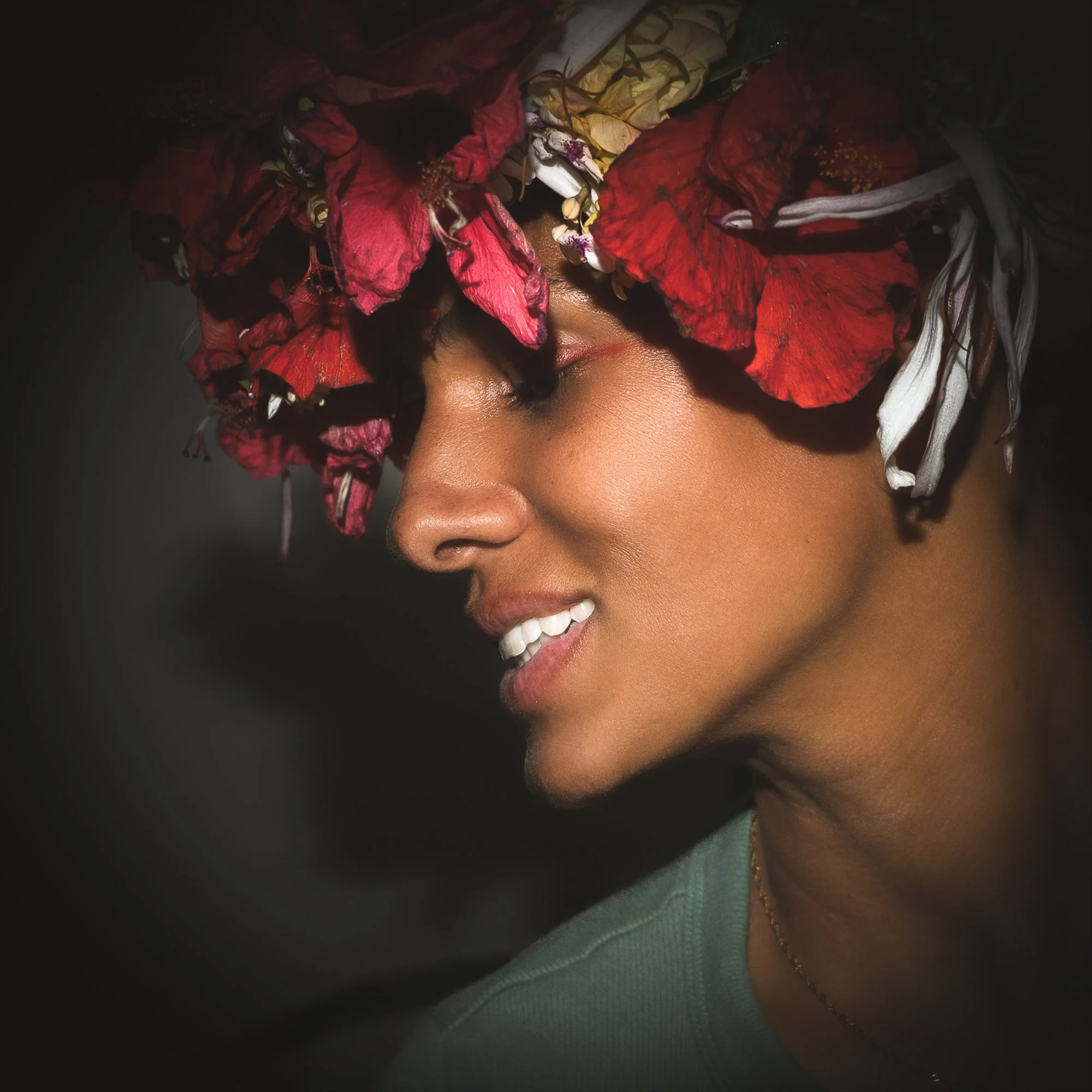 A woman with smooth skin and a bright smile, wearing a headpiece made of vibrant red, pink, white, and yellow flowers, with her eyes closed and in profile.