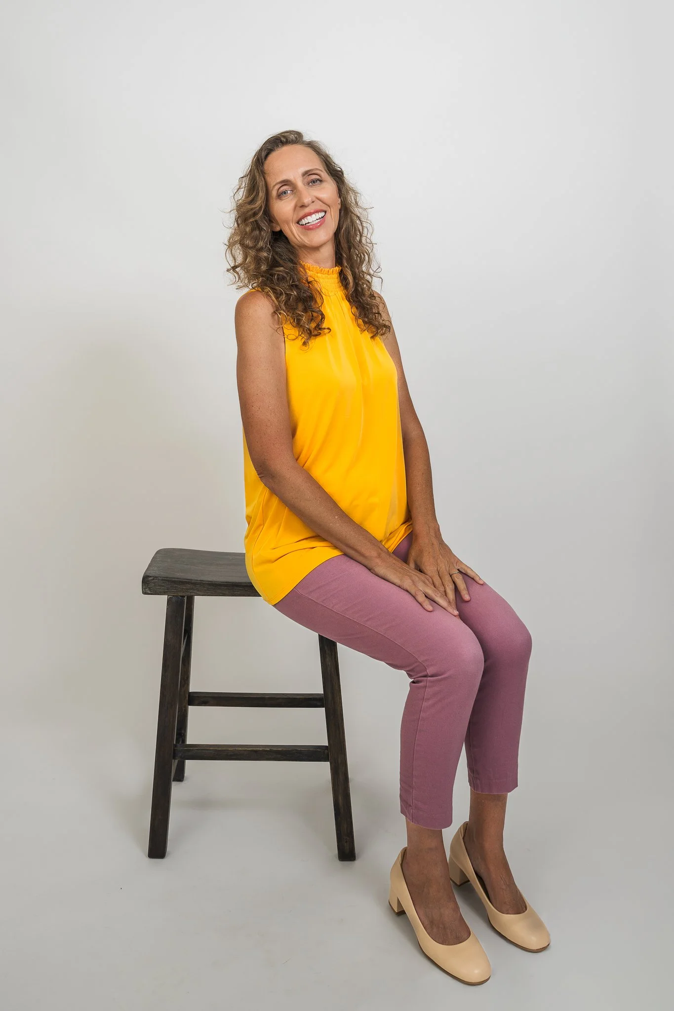 A woman with curly hair, wearing a yellow sleeveless top, pink pants, and beige high heels, sitting on a wooden stool against a plain white background, smiling at the camera.