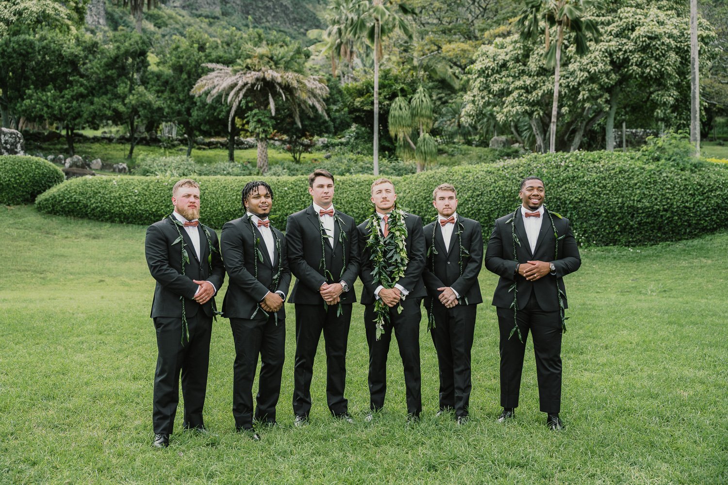 Group of six men dressed in black tuxedos with bow ties, standing on a well-manicured lawn in a lush garden with trees and bushes in the background.