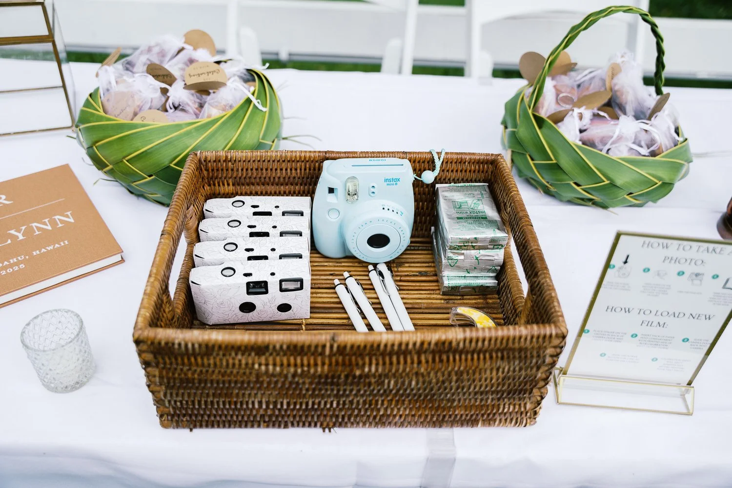Table with a wicker basket containing a blue instant camera, white pens, and film, flanked by two green woven baskets with gifts wrapped in white paper and ribbons, nearby a glass, an open book, and a sign with instructions for taking photos and load