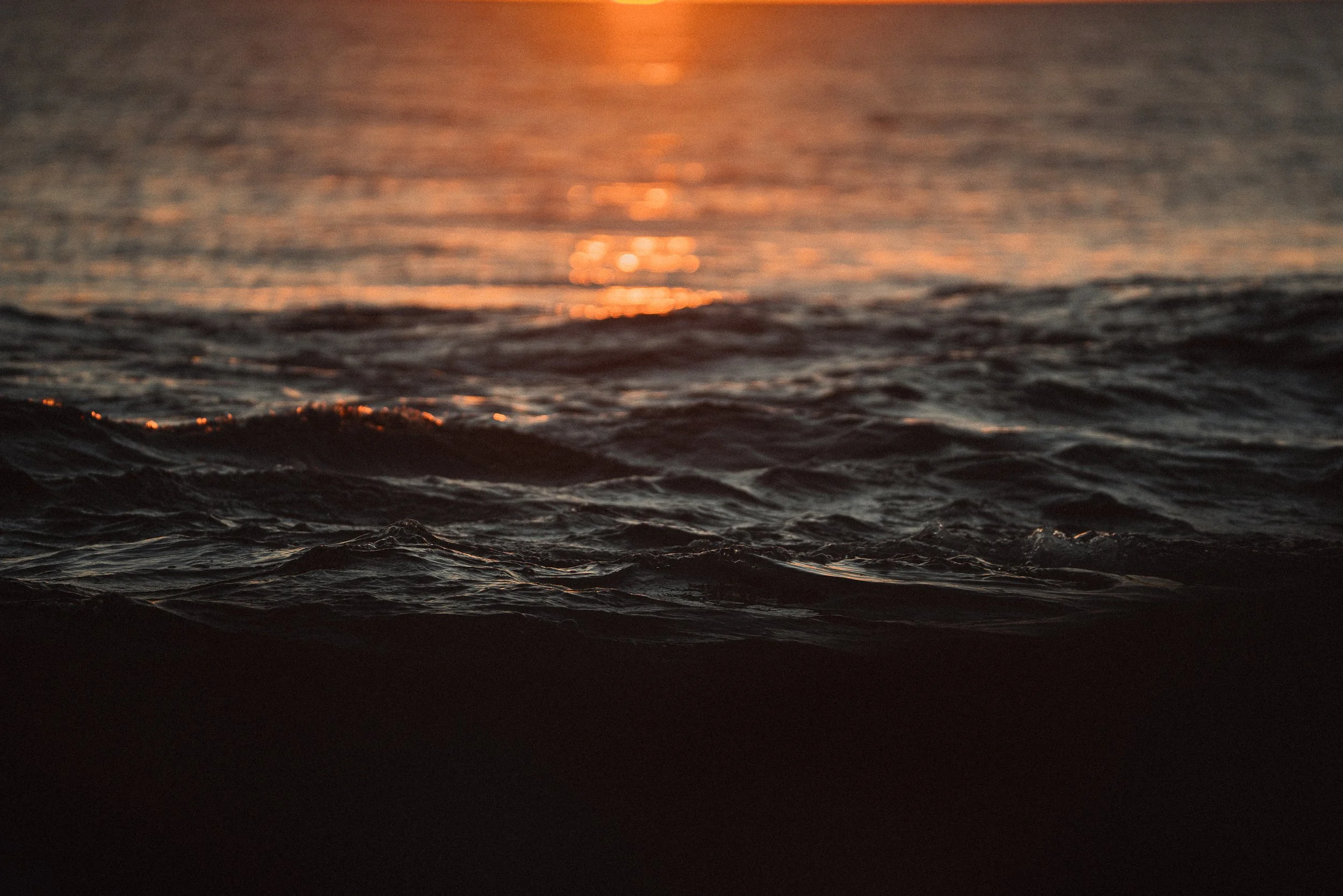 Close-up of ocean waves at sunset with orange glow on horizon.