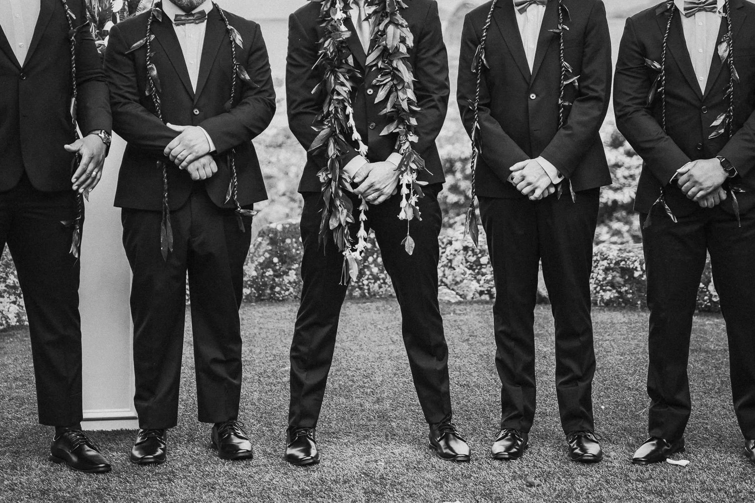A group of men dressed in black suits, some with leis, standing outside on a gravel surface with flowers in the background, all photographing or attending a formal event.