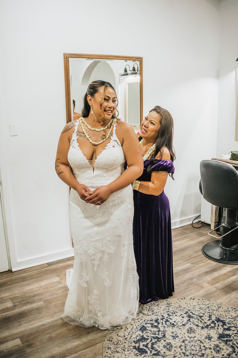 A bride in a white lace wedding dress smiling and looking away as an older woman, dressed in a purple gown, adjusts her dress and touches her shoulder, both standing in a room with white walls and a mirror.