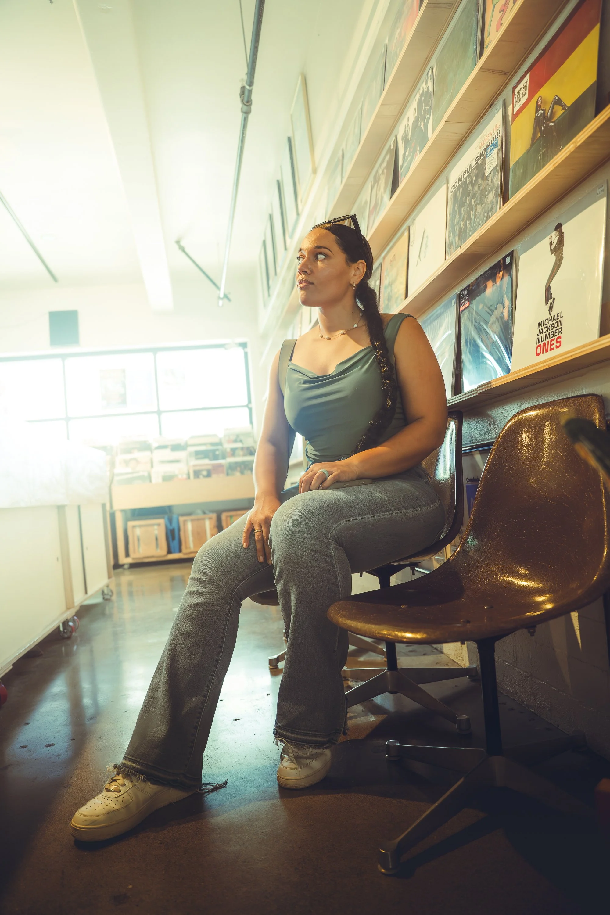 A woman sitting in a vintage-style chair in a recording or music store, looking sideways with bookshelves and vinyl records visible in the background.