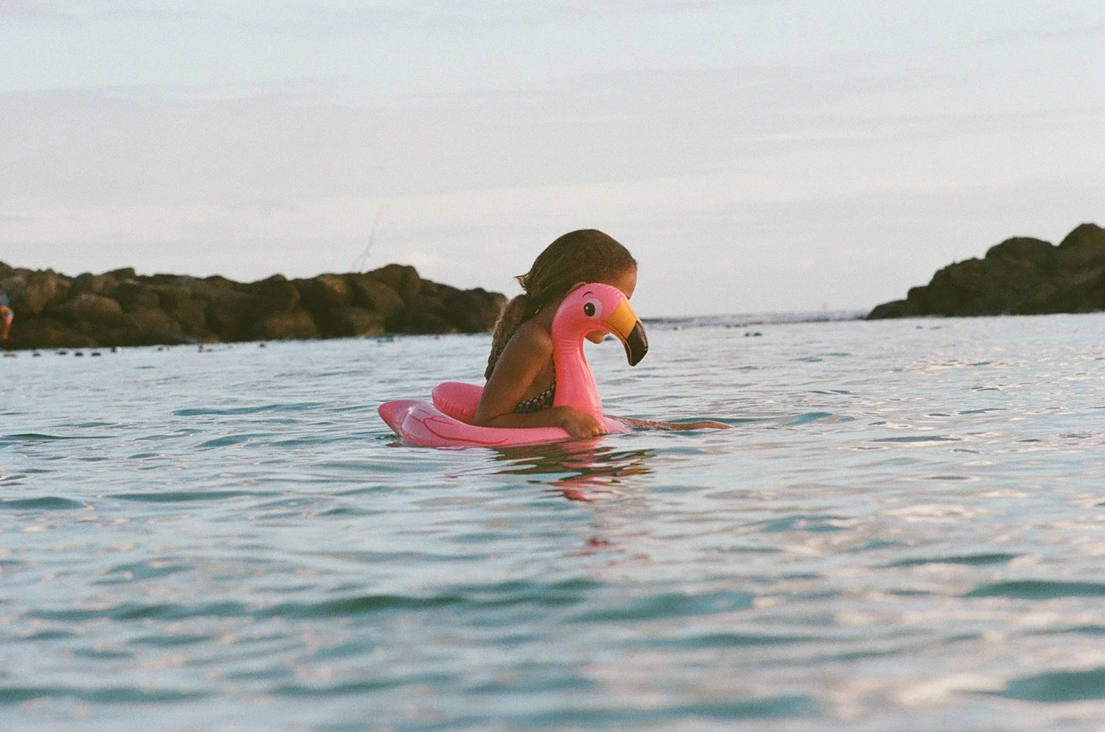 Child with braids wearing a swimsuit, sitting on a pink flamingo float in the water at the beach, with rocks in the background.