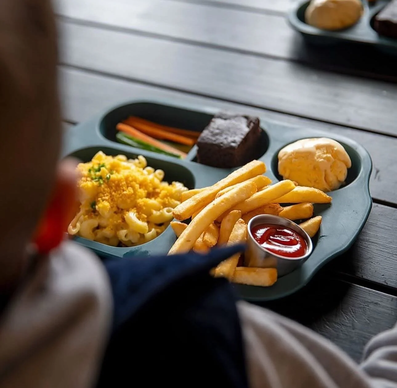 A plate of food containing macaroni and cheese, French fries with ketchup, a scoop of mashed potatoes, a piece of chocolate cake, and some sliced vegetables on a wooden table.
