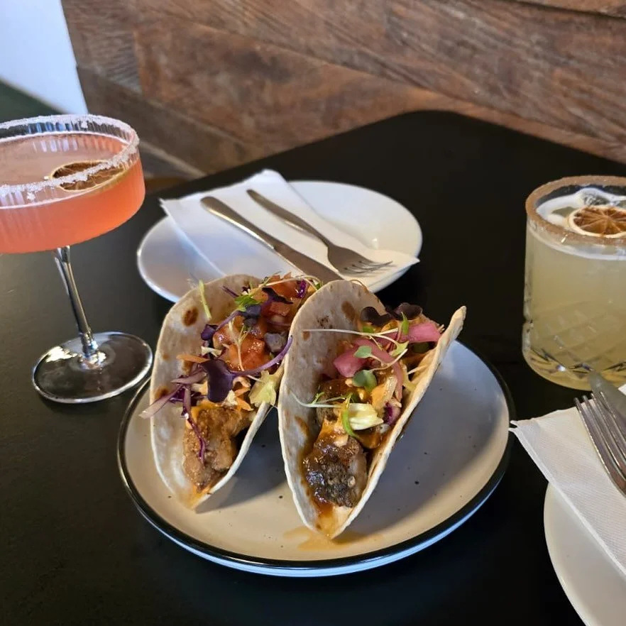 Two tacos with meat, vegetables, and garnishes on a white plate, two cocktails, and utensils on a black table.