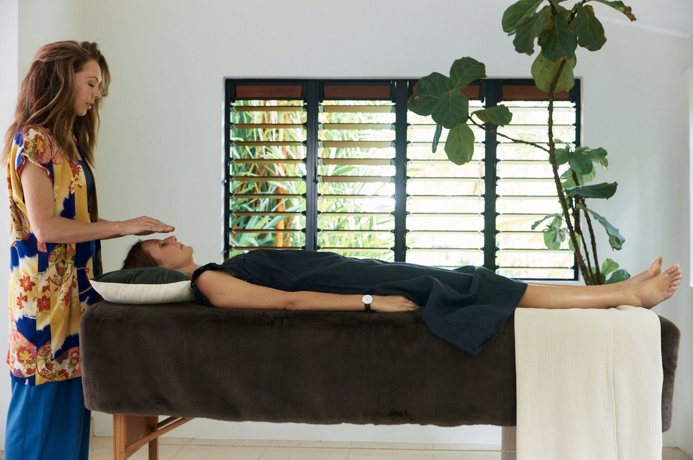 Woman lying on a therapy table with eyes closed, another woman standing beside, gently touching her forehead, inside a bright room with large window and green plants.