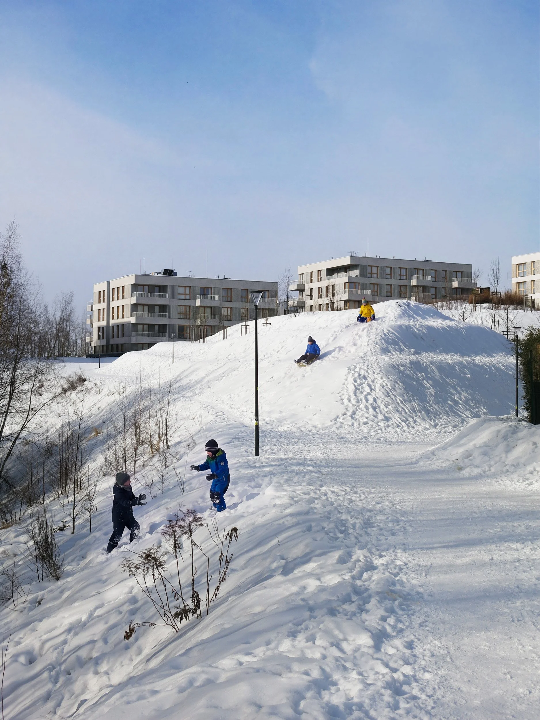 A winter scene showing children playing on a snow-covered hill in a modern residential area. Contemporary apartment buildings are visible in the background, contrasting with the natural, snowy landscape. The image captures the atmosphere of outdoor a