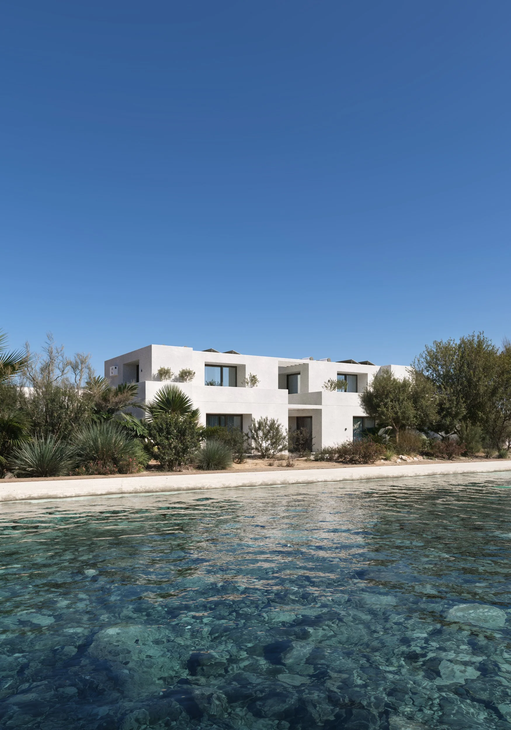 Modern greek Mediterranean-style villa by crystal-clear water. White minimalist architecture surrounded by exotic greenery and a bright blue sky creates a luxurious seaside atmosphere. Kamil Rombczyk portfolio architectural vizualization. Kamil Rombc