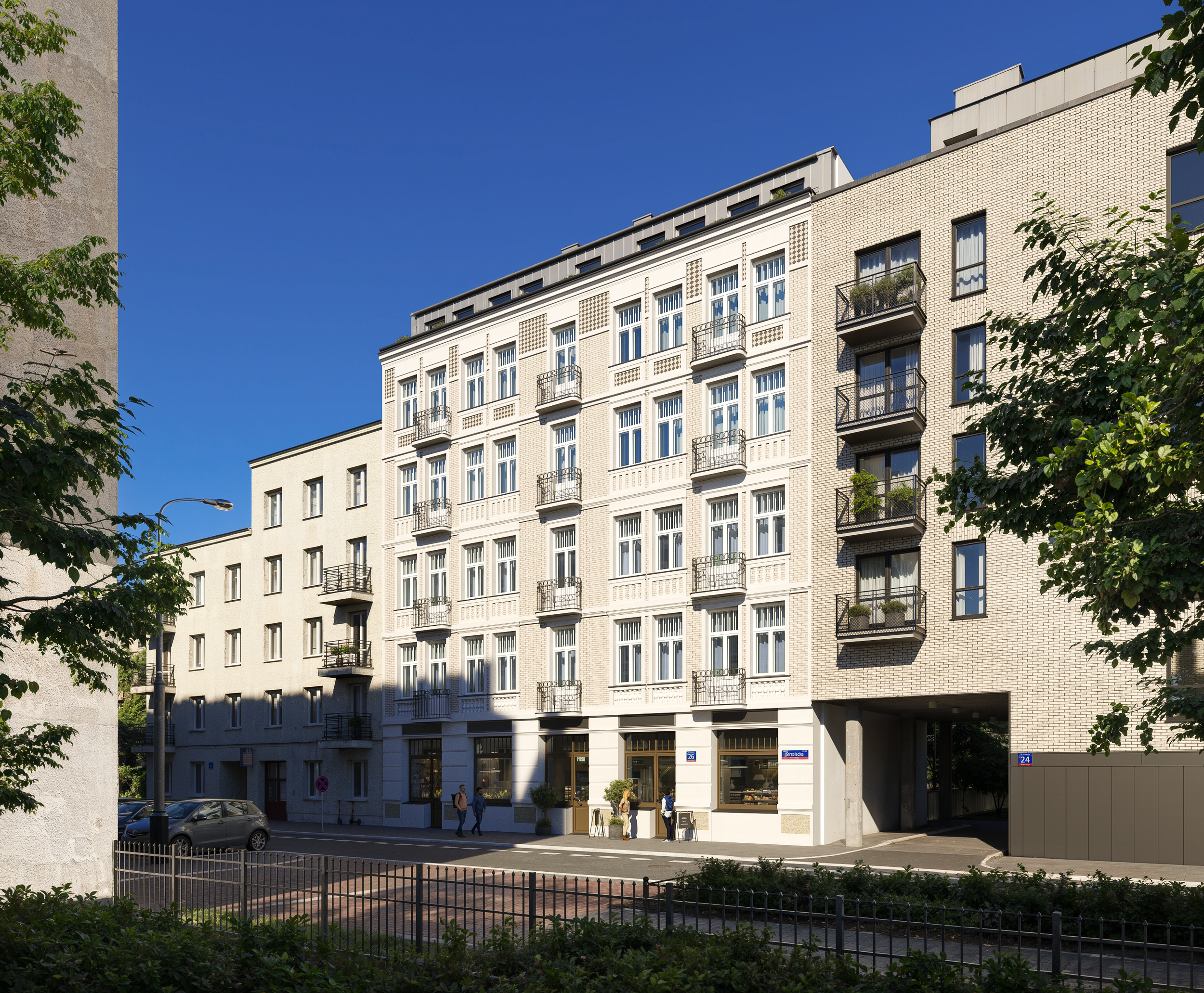 This image presents a stylish urban residential building in Warszawa - kamienica. with elegant light-brick façades, classic balconies, and large windows that create a bright, welcoming appearance. The ground floor features commercial spaces, adding v