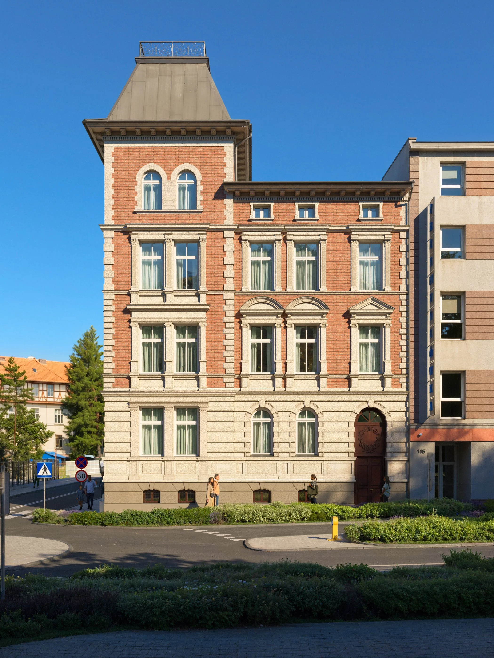 A multi-story building with a combination of red brick and beige stone exterior, tall narrow windows, and a distinctive tower-like upper section topped with a small balcony, situated on a city street with pedestrians and greenery. Kamil Rombczyk port