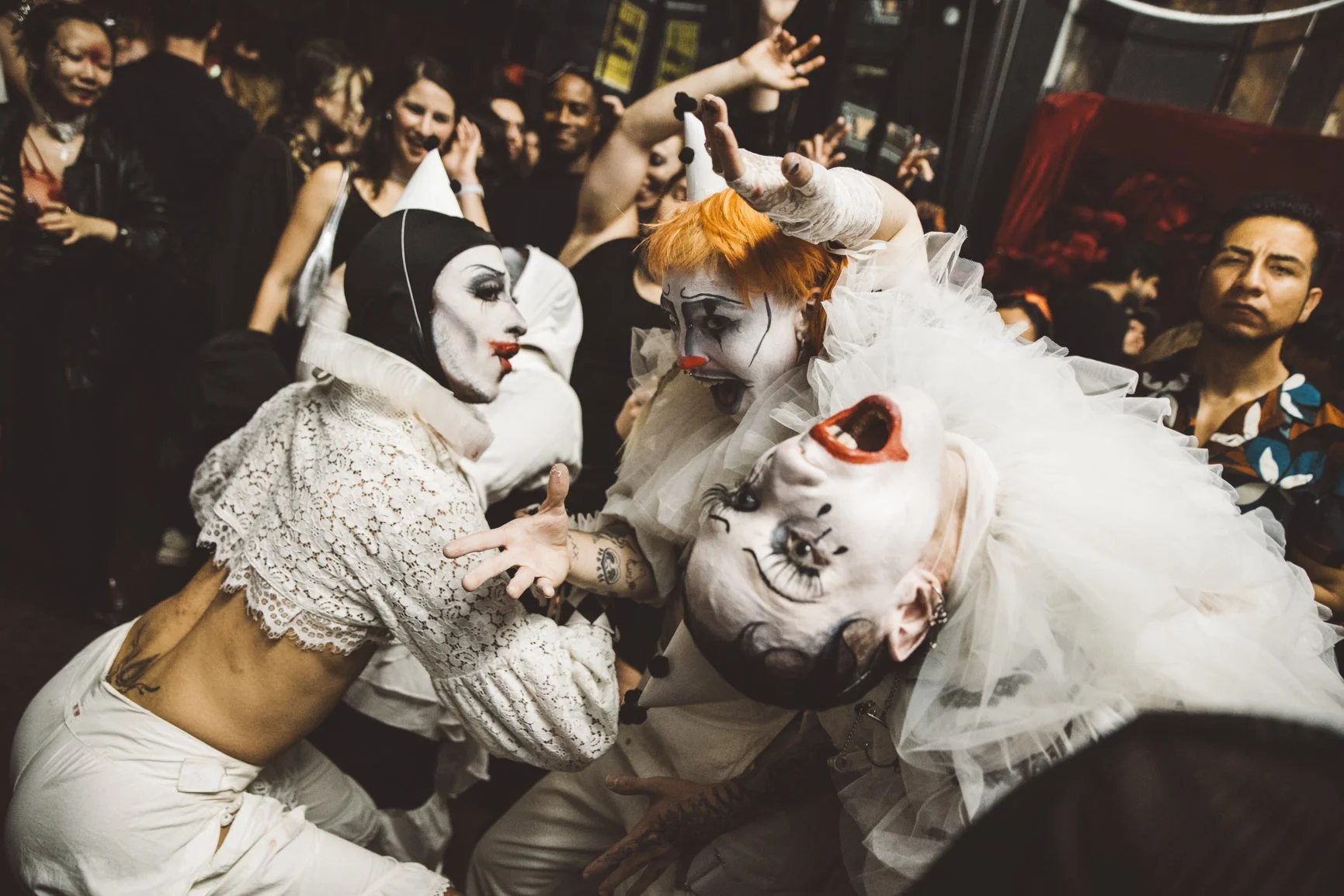 People in clown costumes, some with painted faces, in a crowded indoor party setting.