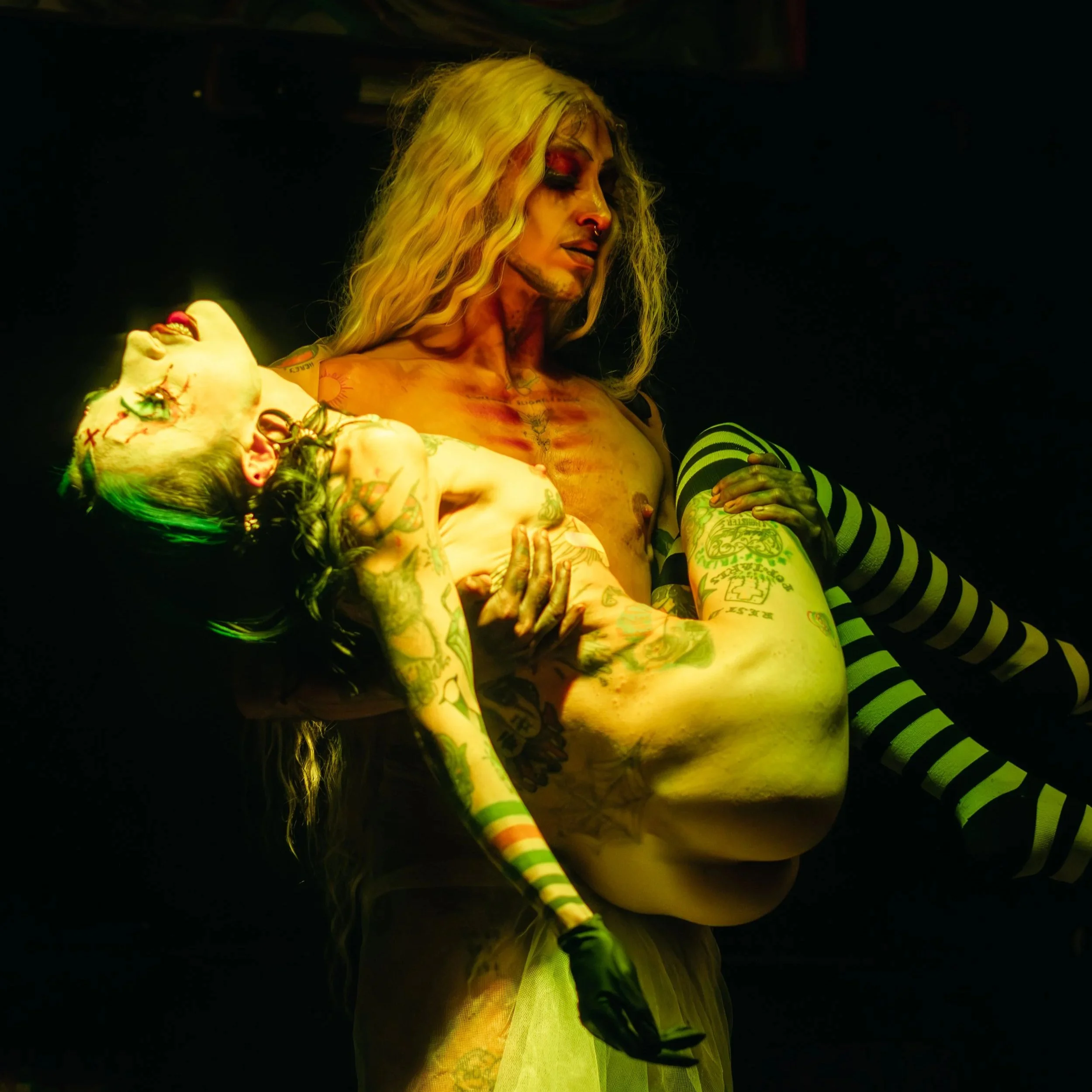 A person with blonde hair holding a heavily tattooed and painted woman, both with edgy, punk-style makeup, in a dark setting.
