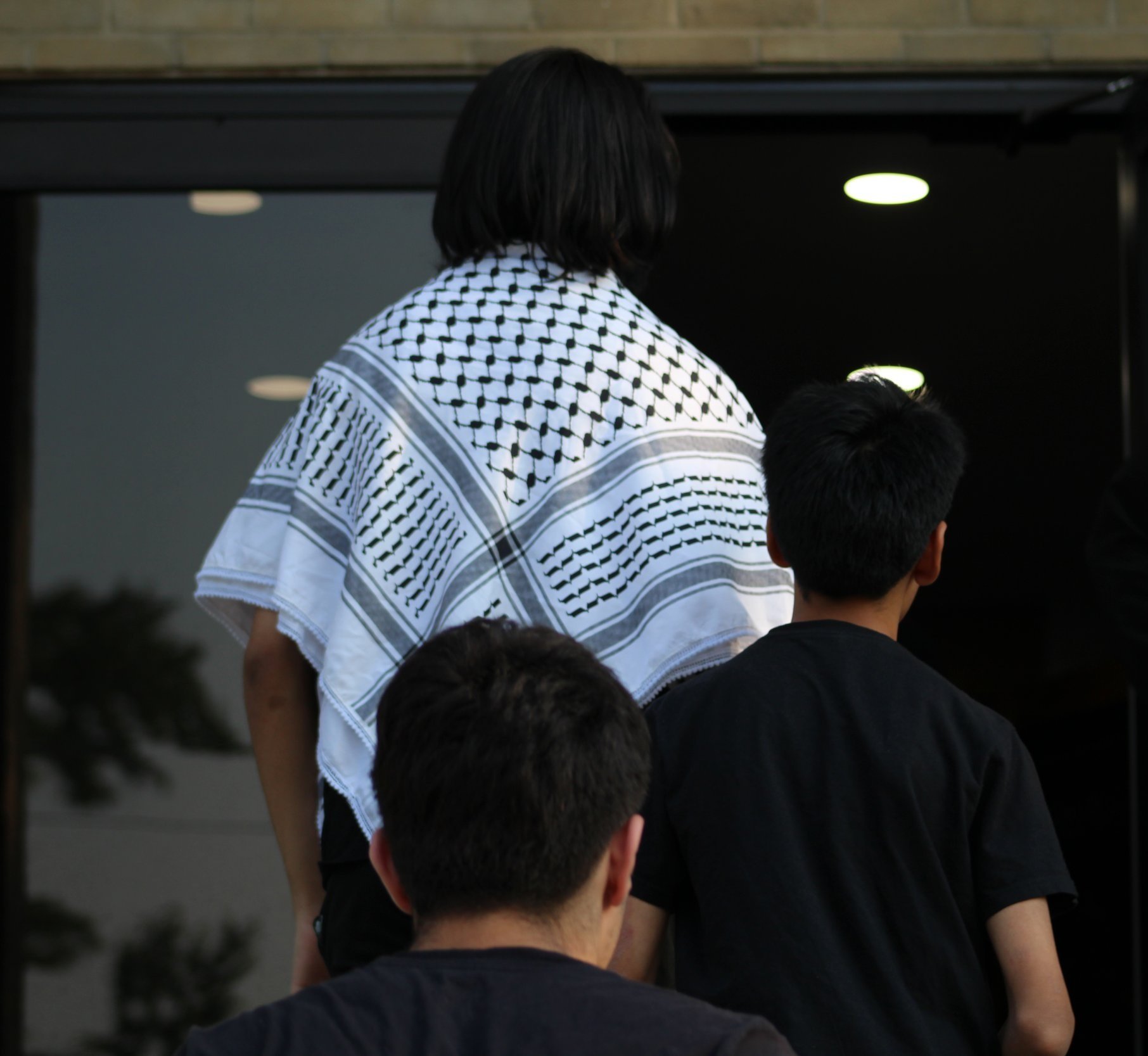 Community members walking into a Light of Guidance lecture program, one wearing a white keffiyeh with a black pattern.