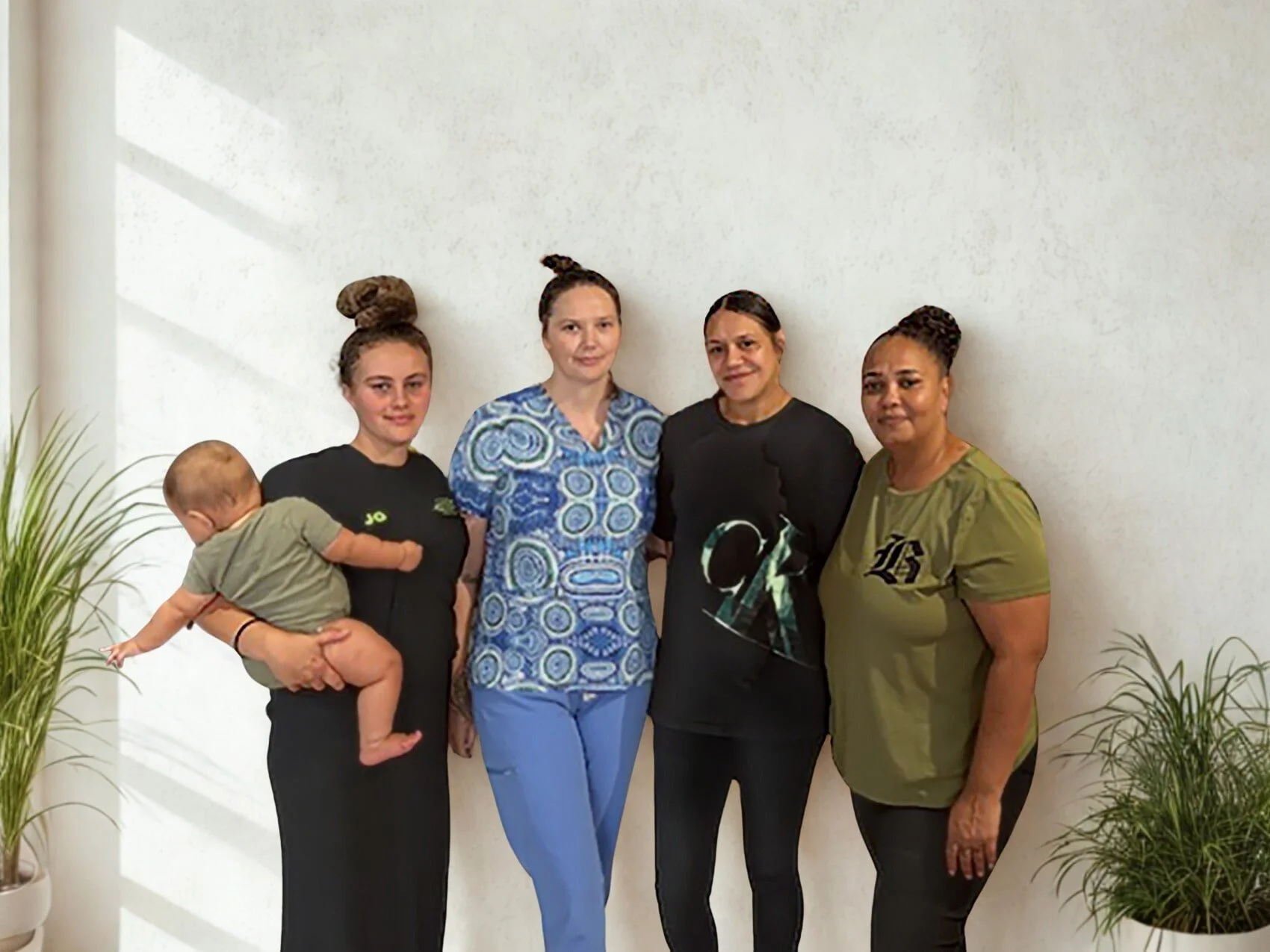 Four women and a baby standing indoors against a white wall, with potted plants on either side.