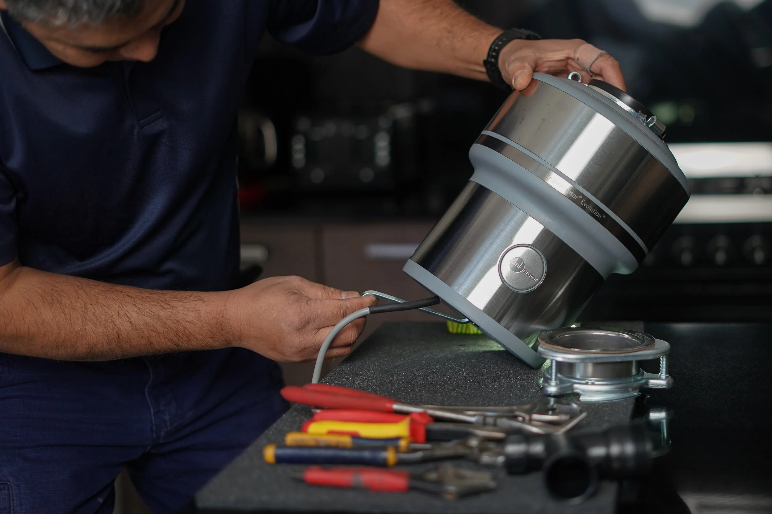 A person installs a garbage disposal unit in a kitchen. Various tools are on the countertop.