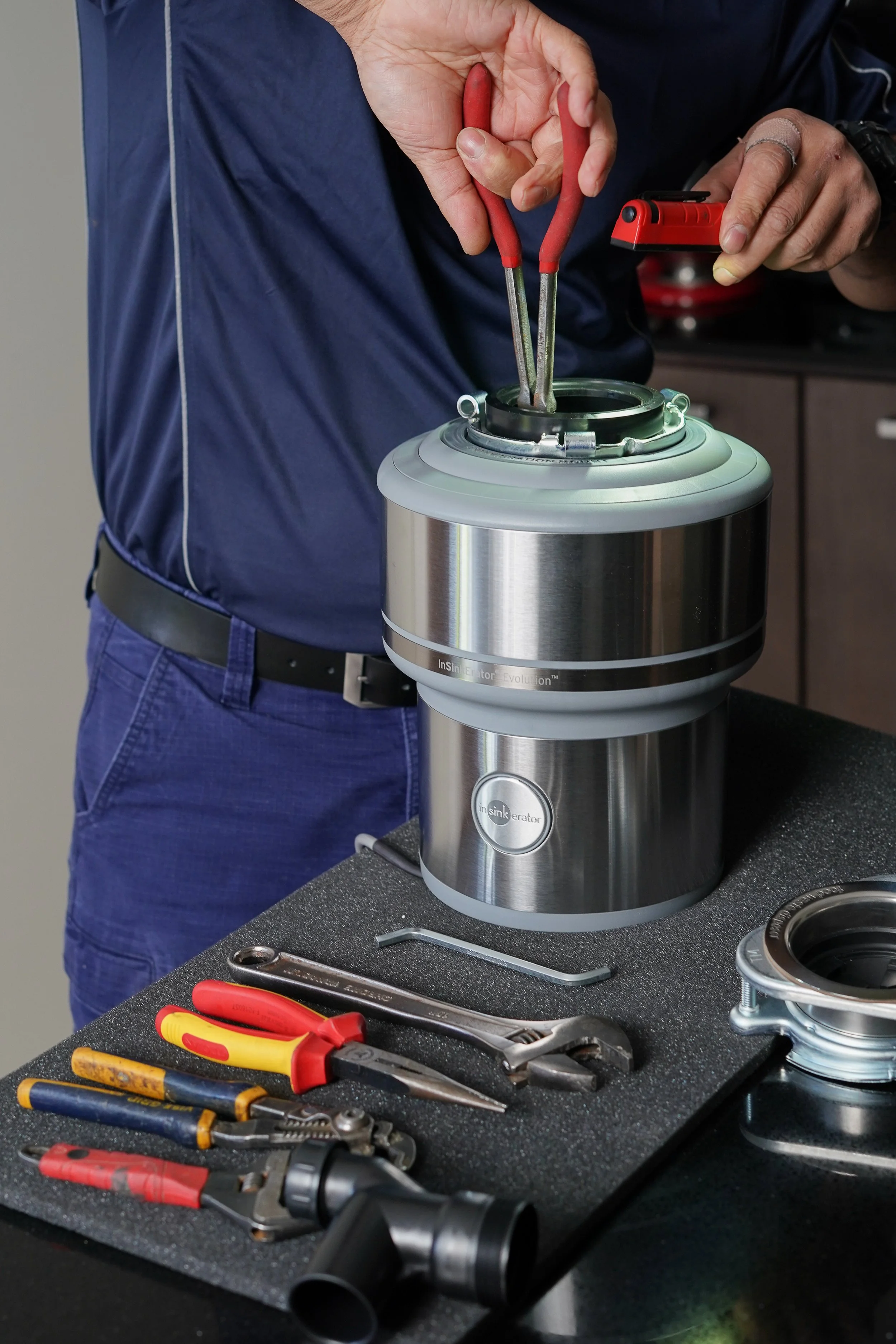 Person repairing a garbage disposal unit with various hand tools on a countertop.