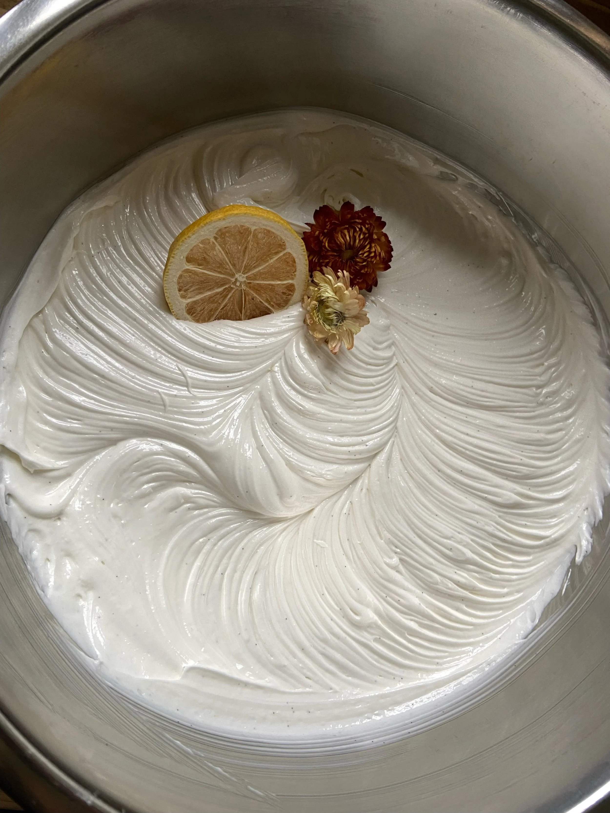 A close-up of a silver bowl filled with thick, swirled white frosting, decorated with a slice of dried lemon and two small dried flowers.