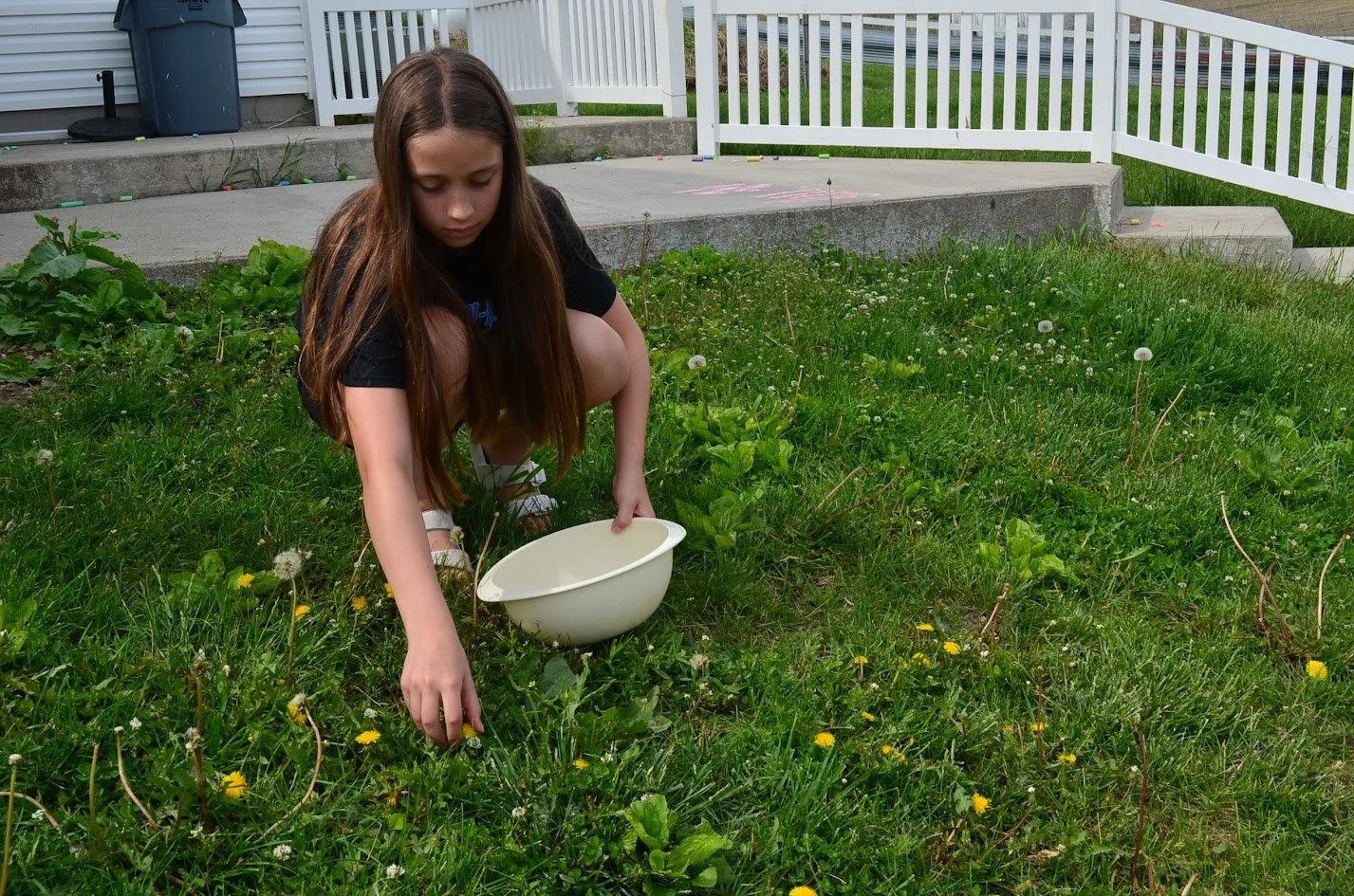 A girl in a black T-shirt, beige shorts, and white sandals crouches on a grassy yard, picking weeds or flowers, with a white bowl nearby. There is a white fence, stairs, and a gray house in the background.