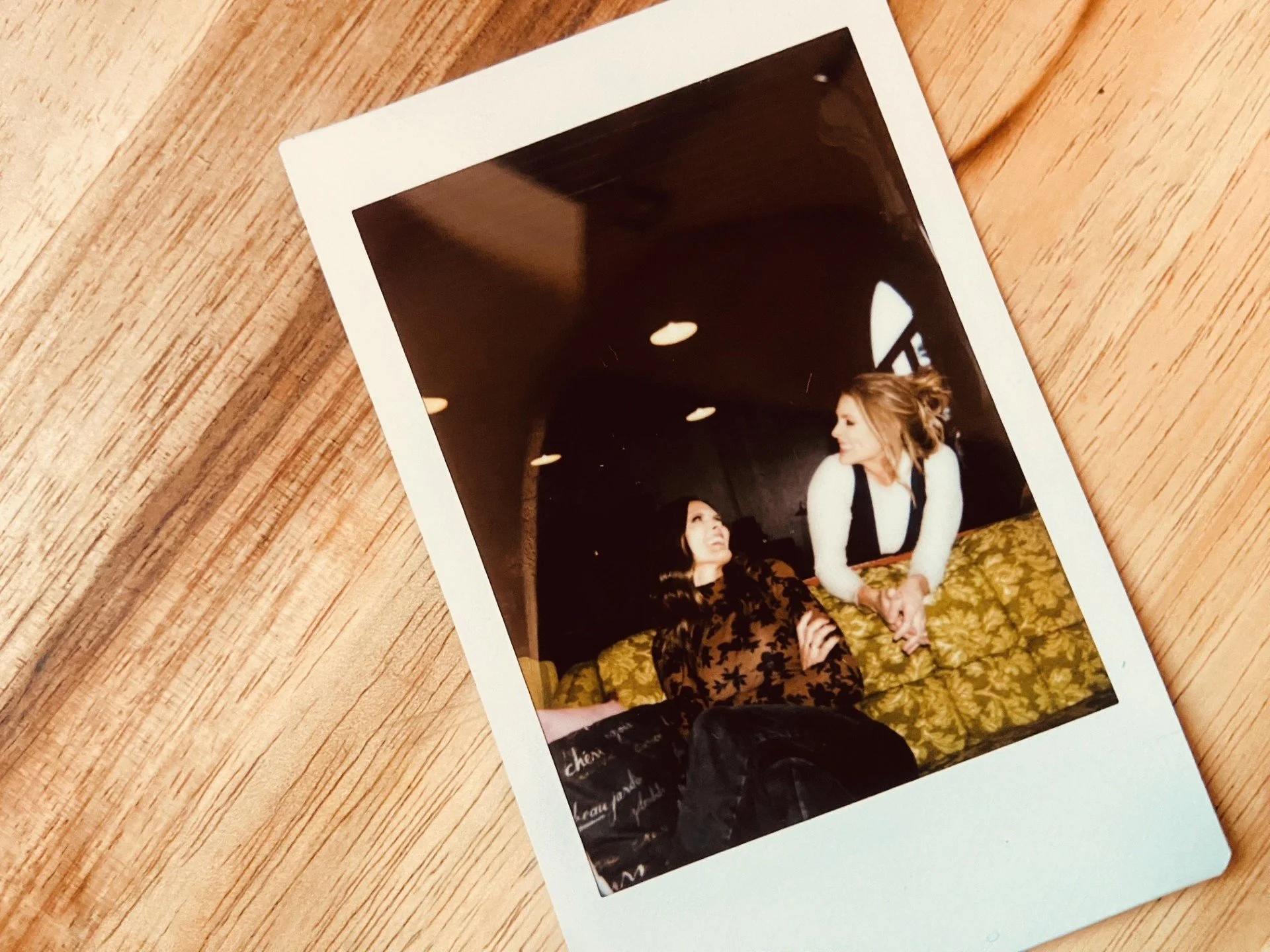 Photograph of two women sitting on a yellow patterned couch engaged in conversation, with one woman listening and the other speaking, in an indoor setting with dark walls and ceiling lights.
