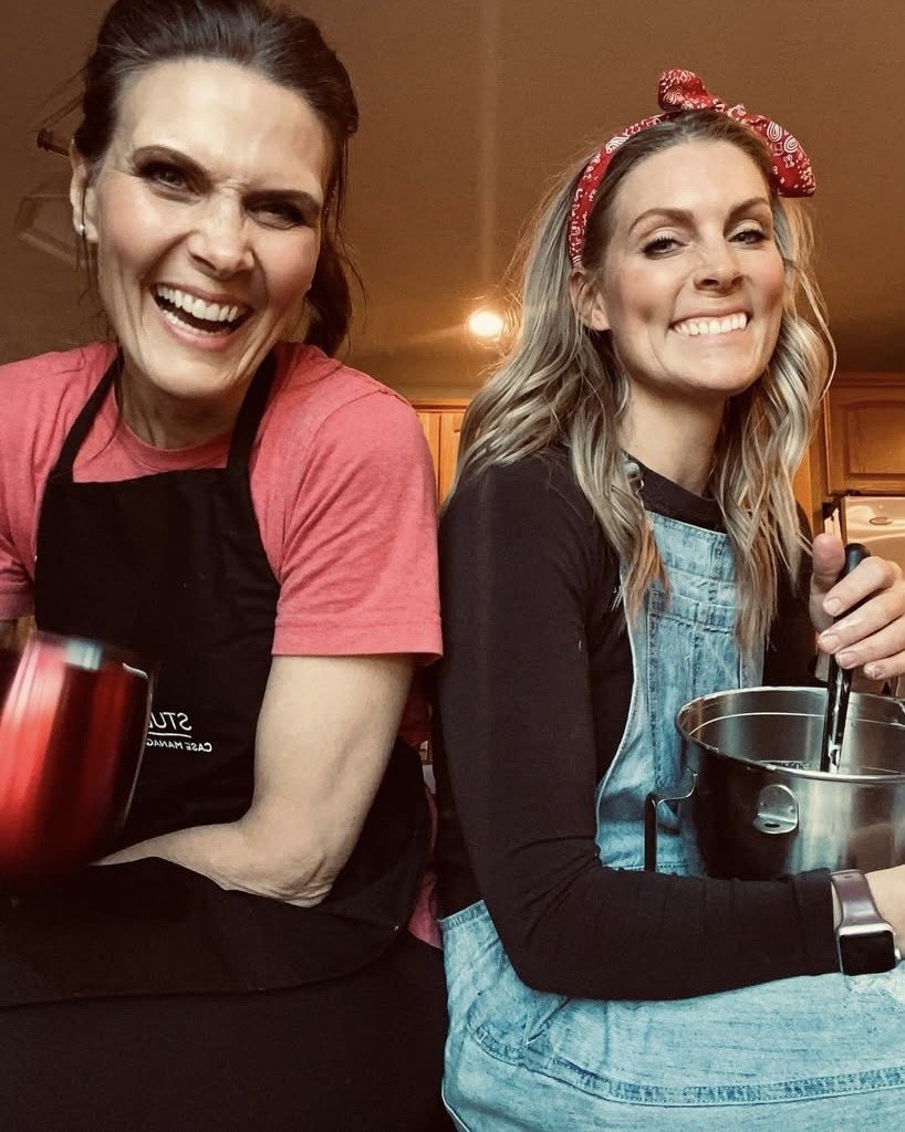 Two women smiling and holding kitchen utensils, one wearing an apron and the other a headband, in a kitchen setting.