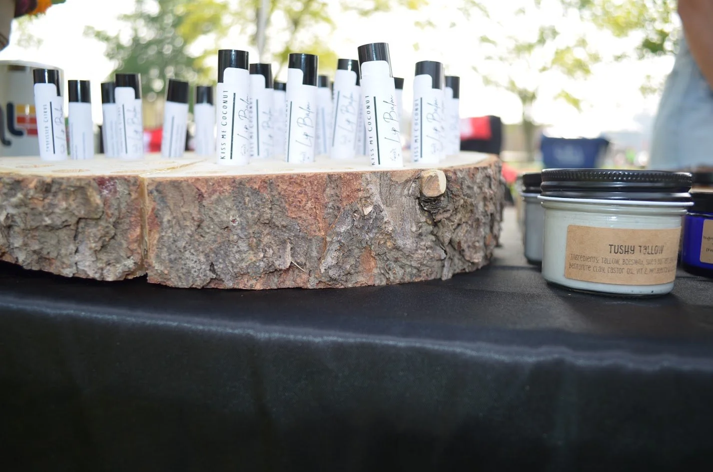 Assorted tallow skincare products on a black table, with a large wood log slice and small jars labeled with natural ingredients like tallow, nestled outdoors under trees.