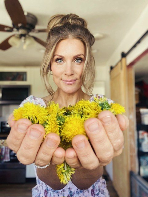 Andrea is holding yellow dandelions and smiling indoors.