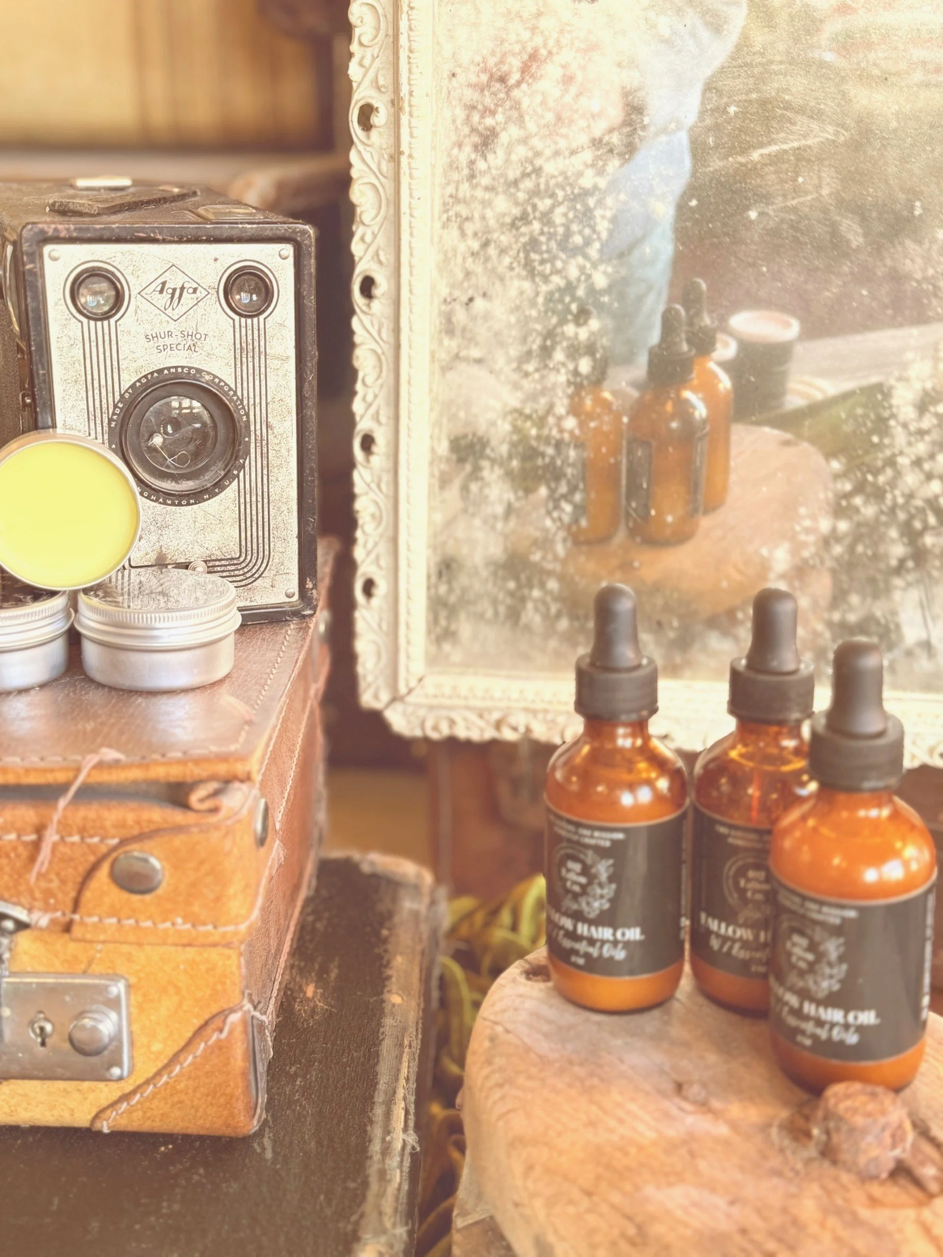 Vintage camera, amber glass dropper bottles labeled as hair oil, and tins on a wooden surface with an ornate mirror reflecting the bottles.
