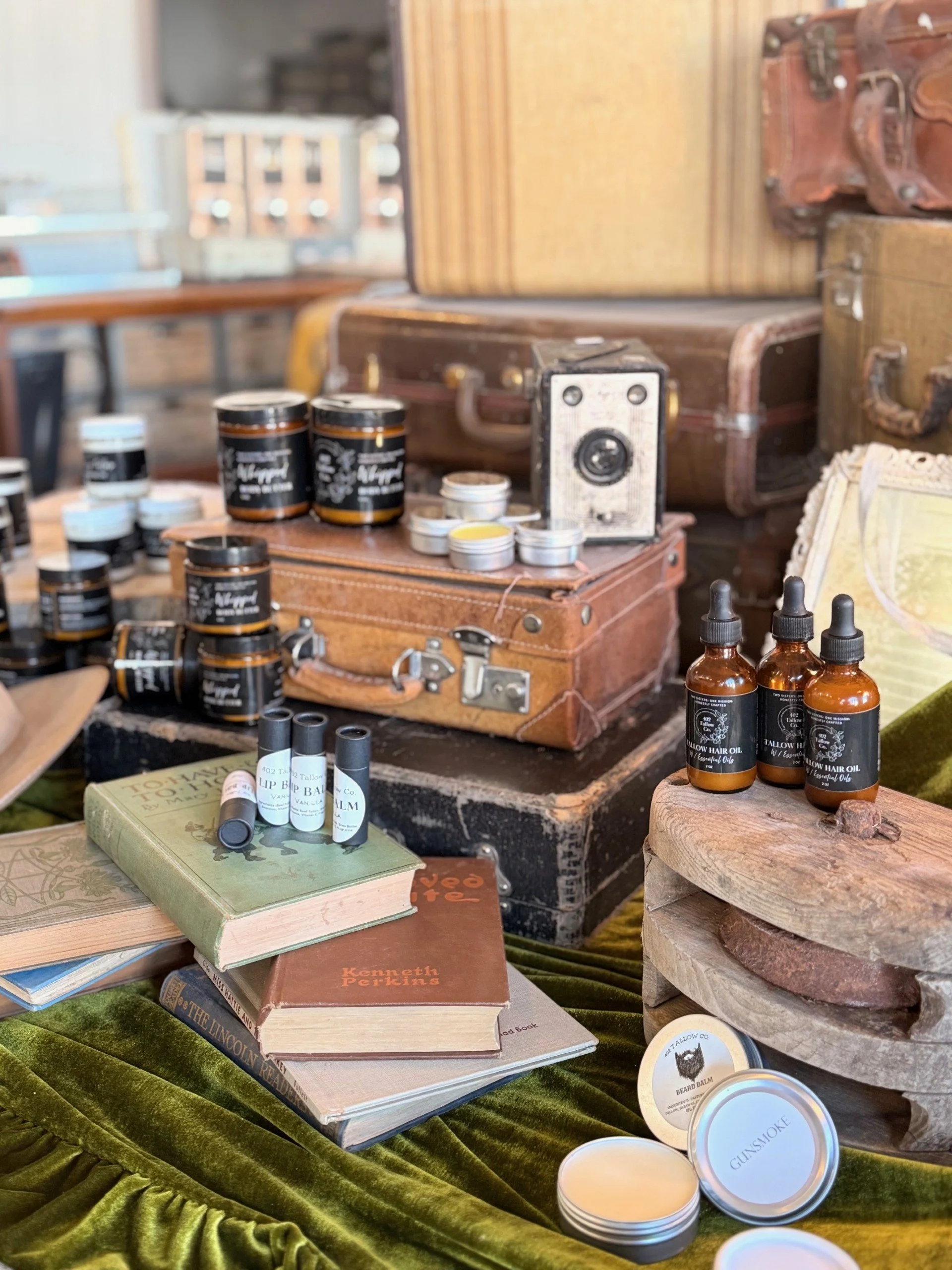 Vintage and rustic items including leather suitcases, small jars, dropper bottles, books, and a wooden stand with skincare or grooming products on a green velvet tablecloth.