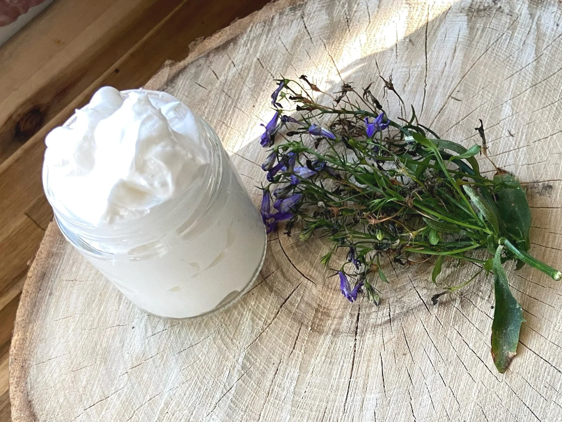 A jar of white cream or yogurt placed next to a bunch of purple flowers with green leaves on a tree stump.