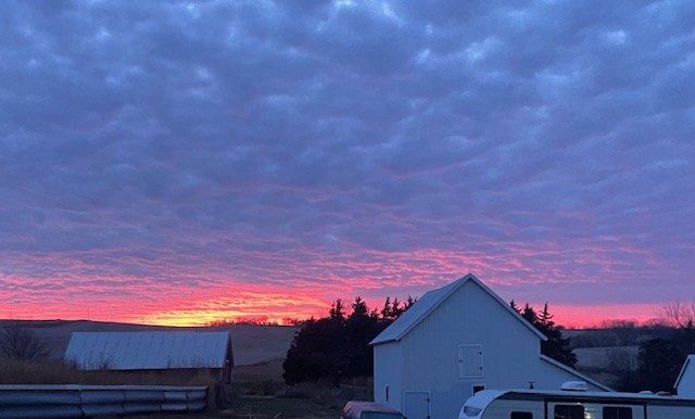 A rural landscape at sunset with a vibrant pink and orange sky, a white barn, trees, and rooftops in the foreground.