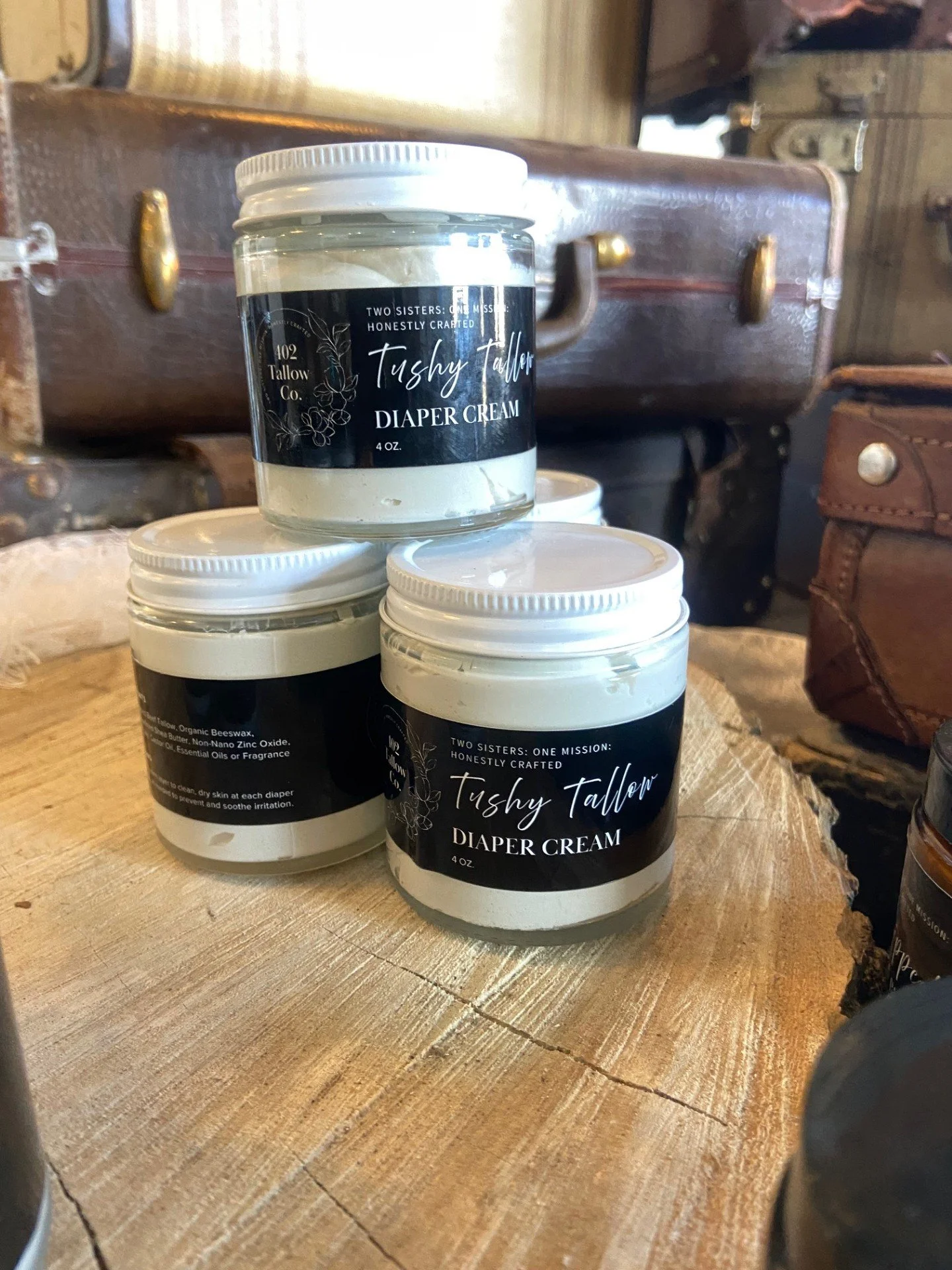 Three jars of diaper cream labeled 'Tushy Tallow Diaper Cream' on a wooden surface, with a vintage suitcase in the background.