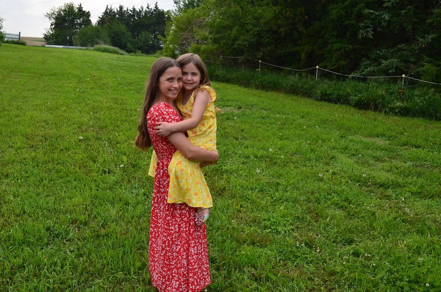 Two girls, one older in a red floral dress holding a younger girl in a yellow floral dress, standing on a grassy field with trees in the background.