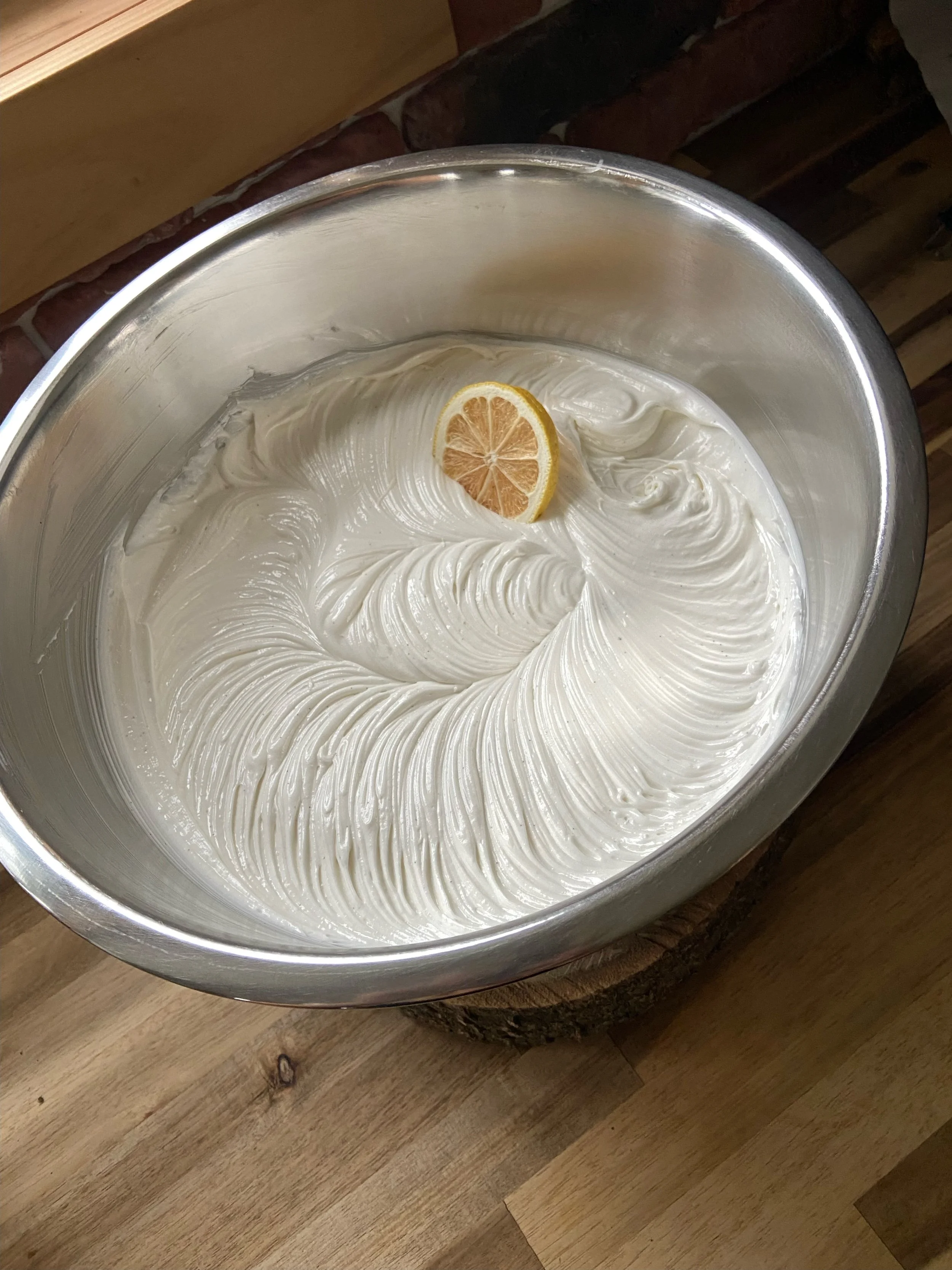 A stainless steel mixing bowl with whipped cream and a lemon slice on top, placed on a wooden surface.