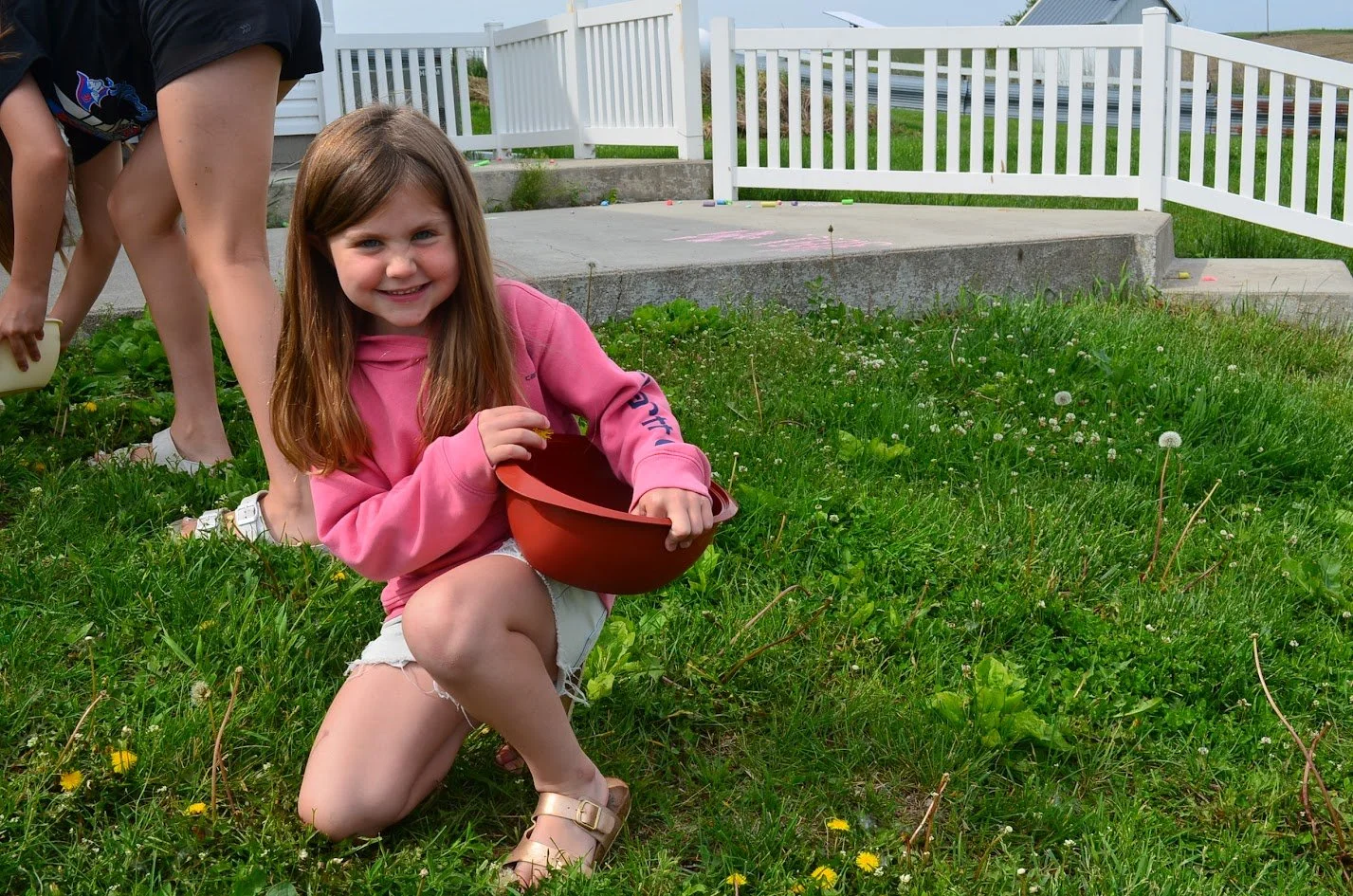 A young girl with long brown hair smiling and kneeling on green grass, holding an orange plastic bowl. She is wearing a pink hoodie, white shorts, and sandals. In the background, there is a white fence and another person partly visible, bending down.